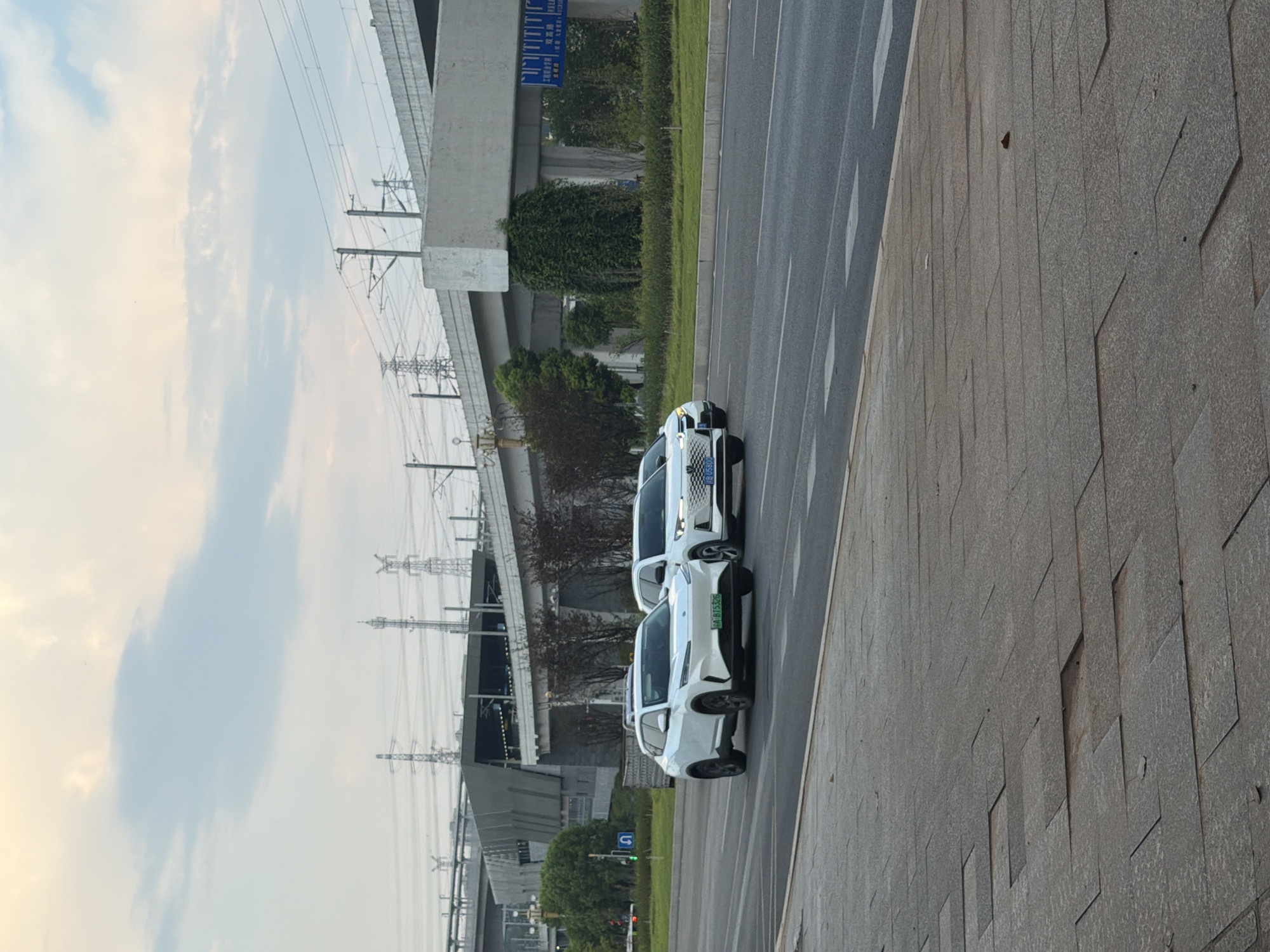 This is a photograph taken in Jiangjin, China, showing a street scene with an elevated highway structure in the background. Two white electric cars are driving on the road. The car in the foreground is a white SUV with a license plate that reads "A BT5326". The second white car, slightly behind and to the right, is also an SUV with a license plate that reads "B U5800". Both cars are moving forward on the asphalt road. The road surface has lane markings. To the left of the road, there is a sidewalk made of large, square paving stones, some of which appear to be uneven or damaged. Green trees and bushes line the area between the road and the elevated highway. The sky is cloudy with hints of blue. Several utility poles with power lines are visible in the distance, as well as parts of the elevated highway structure, which appears to be a train or metro line. A traffic light can be seen on the left side of the road. The overall atmosphere suggests an urban environment during daylight hours.