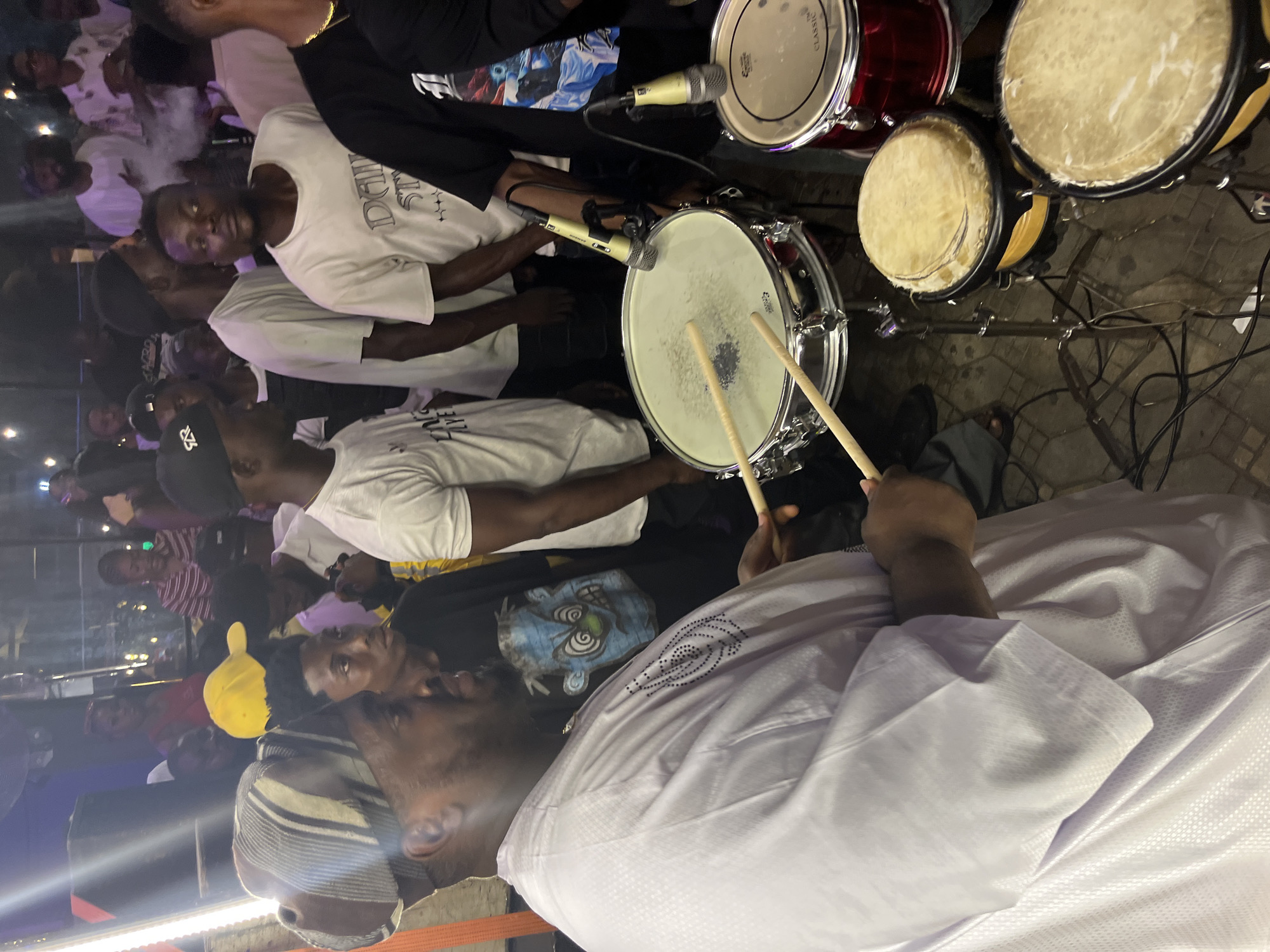 In the city of Unknown, Unknown, a group of people are gathered, likely at an outdoor event or club. In the foreground, a man dressed in a white traditional outfit and a striped cap is playing a drum set. He is holding drumsticks and appears to be in the middle of a performance. The drum set includes a snare drum, a bass drum, and two smaller conga drums. Microphones are positioned near the snare drum, suggesting the performance is being amplified.
Behind the drummer, other individuals are visible, some of whom are also dressed in casual attire. Some are observing the performance, while others seem to be engaged in conversation or enjoying the atmosphere. The background is dimly lit, with some lights and smoke contributing to the ambiance. The overall scene suggests a lively and social gathering with a musical element.