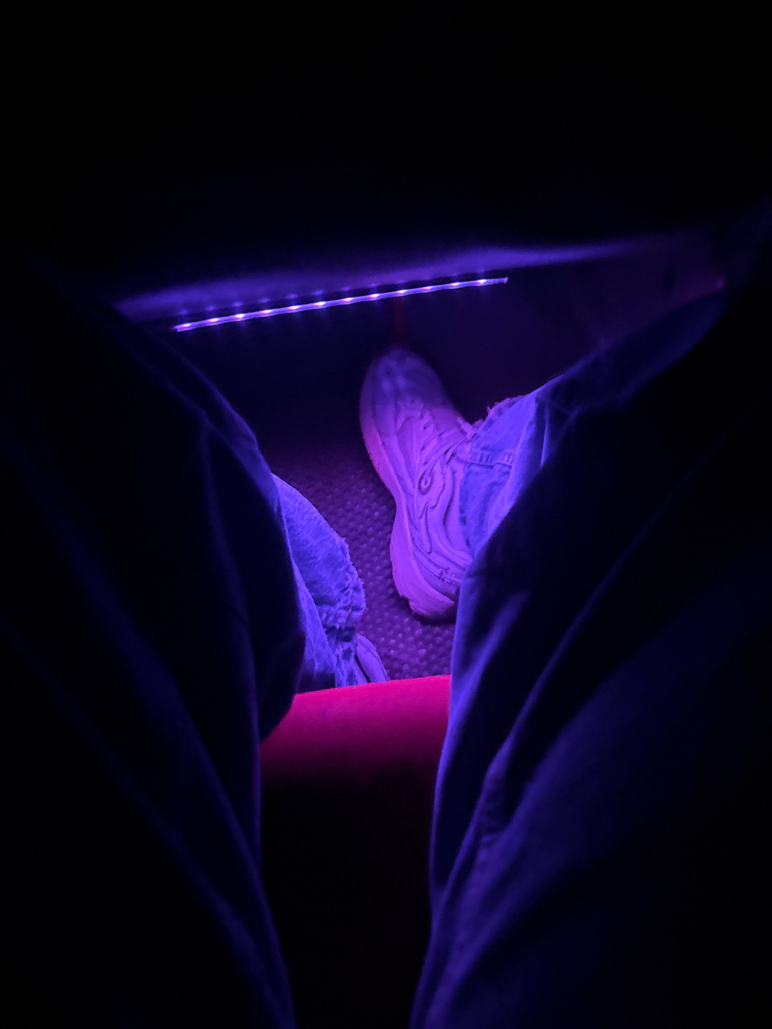 An individual is seated, with their legs extended forward. One foot, clad in a light-colored sneaker, is visible. The sneaker appears to be a chunky, athletic style, illuminated by a strip of purple LED lights. The individual is wearing blue denim jeans, which are partially obscured by dark blue fabric, possibly a jacket or blanket. The lighting suggests the setting may be indoors, possibly a vehicle or a room with ambient mood lighting. The texture of the floor beneath the sneaker appears to be a patterned surface, possibly carpet or rubber matting. No specific location in Нарацкі сельскі Савет, Belarus can be determined from the visual information.