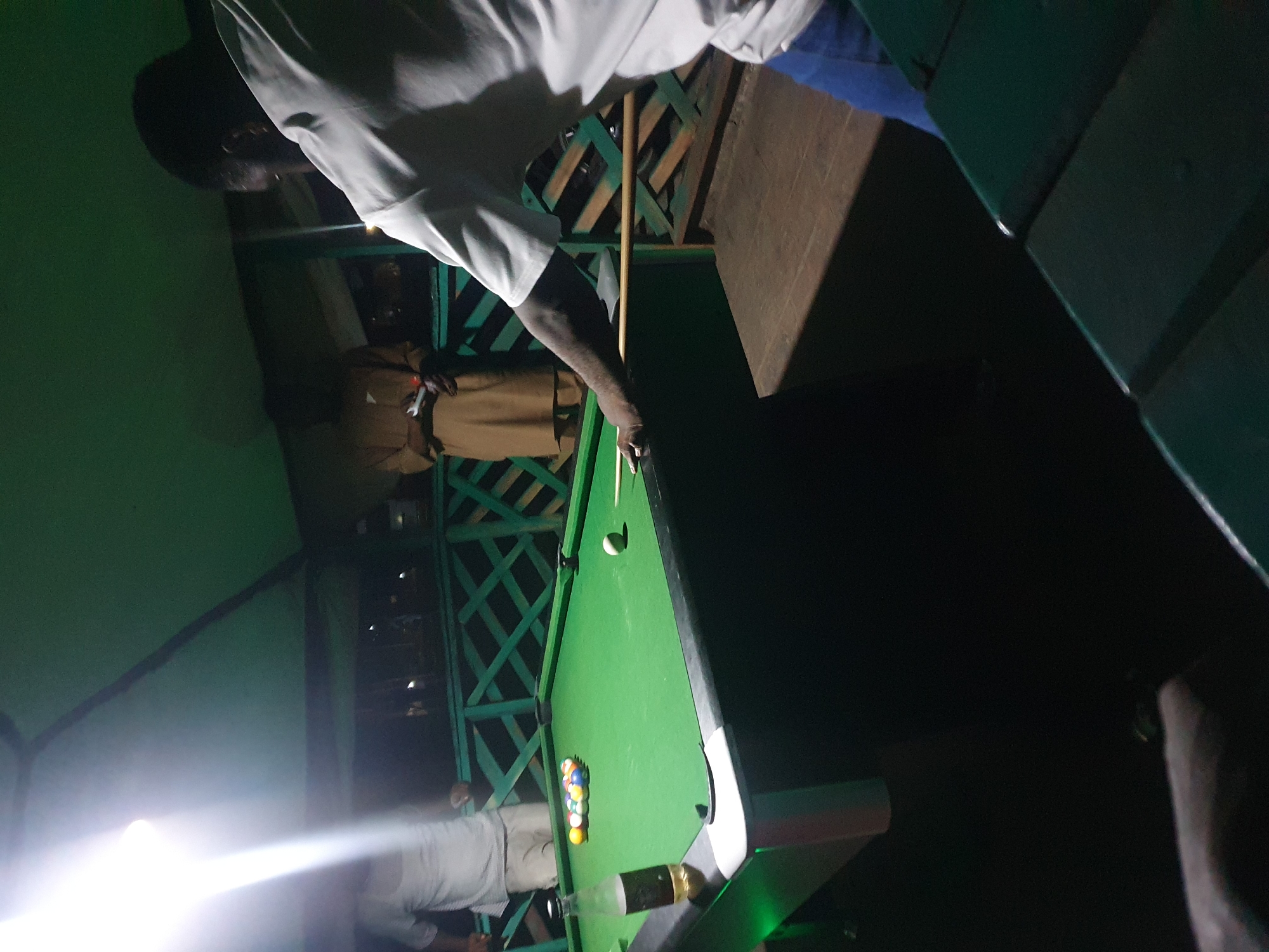 The image depicts an outdoor pool table area at night, possibly at a bar or entertainment establishment in Lokoja, Nigeria. A group of individuals are present. In the foreground, a person in a white t-shirt and blue jeans is leaning over the pool table, holding a pool cue and preparing to take a shot. The pool table is green, with a rack of colored billiard balls on one side and a single white ball on the other. A bottle of what appears to be a beverage is also on the table. In the background, two other individuals are visible. One is wearing a light-colored t-shirt and pants, and the other is wearing a tan robe-like garment. The area is illuminated by overhead lights, casting bright reflections and creating shadows. A green railing surrounds the elevated platform where the pool table is situated. The overall atmosphere suggests a casual, social gathering.