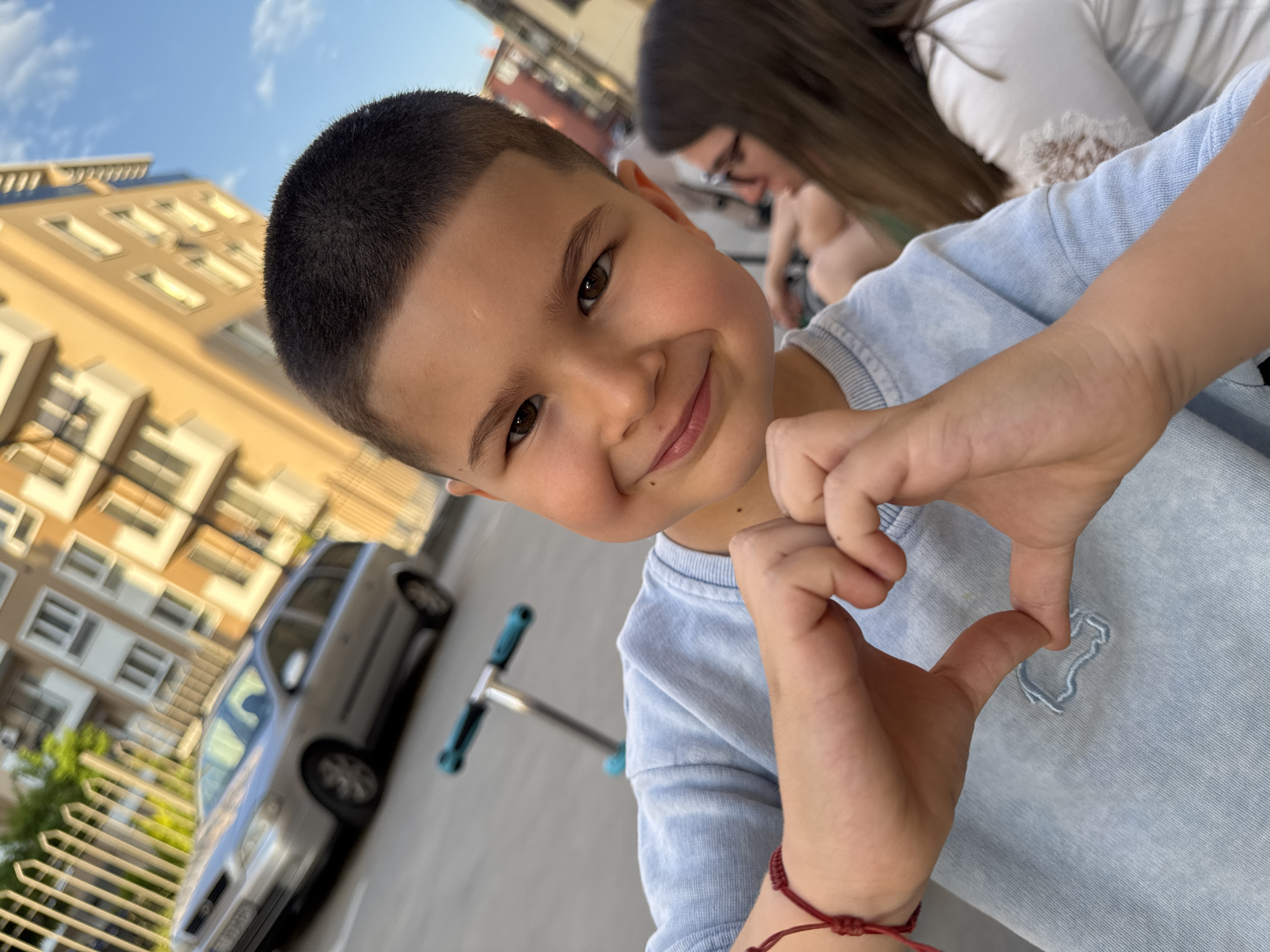 A smiling young boy with a close-cropped haircut is making a heart shape with his hands. He is wearing a light blue t-shirt. A dark red bracelet is visible on his wrist.
In the background, there is a silver car and a blue scooter. A building with multiple windows is visible, and a woman with glasses can be seen partially in the upper right corner.
Given the architectural style of the building in the background, it is possible this image was taken in a neighborhood in Plovdiv, Bulgaria. More specific details about the exact location would require further visual information.