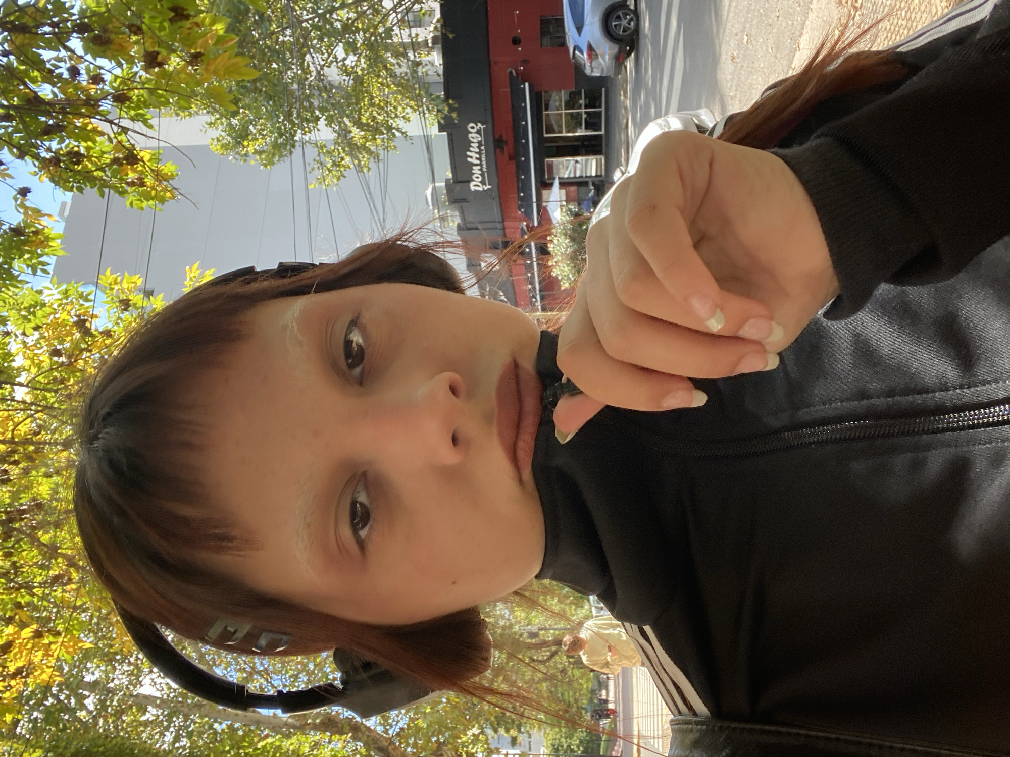 A selfie shows a light-skinned person with light blonde eyebrows and reddish-brown hair, wearing headphones, a black jacket with white stripes, and standing outside. In the background is a tree, the corner of a building, and a sign that reads "Don Hugo Parrilla," indicating that the location is Buenos Aires, Argentina.