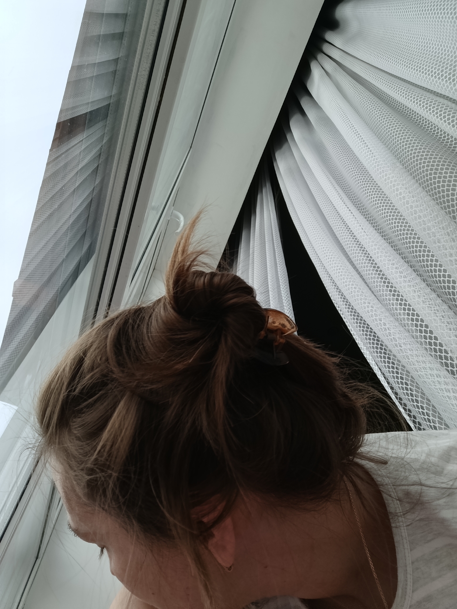 A woman is looking out of a window. The window has a blind and a thin, white curtain. The woman has brown hair in a bun held by a brown clip. A gold chain is visible on her neck. The location is Chaikovsky, Russia.