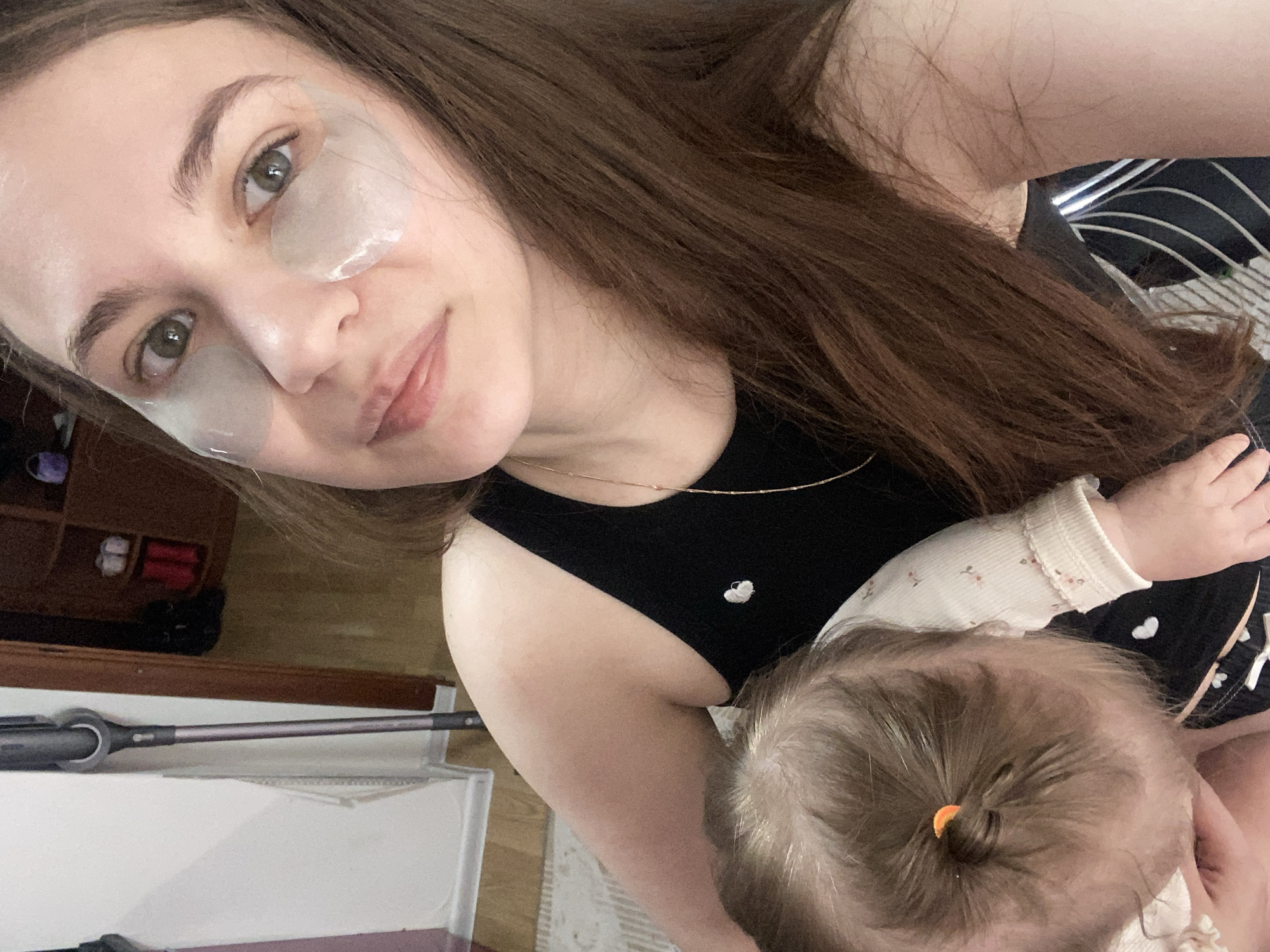 The image displays an adult female holding an infant or toddler in an indoor domestic setting. The adult, with long brown hair, green eyes, and wearing a black tank top with a white heart embellishment and a thin gold necklace, has clear gel patches applied under both eyes and looks directly at the camera with a slight smile. A small area of redness is visible on her right upper arm. The infant or toddler is partially visible in the lower left, with light brown hair tied with a small orange elastic, wearing a white long-sleeve top and dark bottoms with a white pattern, facing away from the camera.
In the background, a dark grey cordless stick vacuum cleaner leans against a light-colored wall to the left. Through an open dark wooden doorway, light wooden flooring and a brown wooden multi-shelf unit with various items, including red footwear, are visible. A cream-colored patterned rug is present on the floor in the foreground. The specific location within the city of Chaikovsky, Russia, cannot be determined from the visual evidence provided in the image.