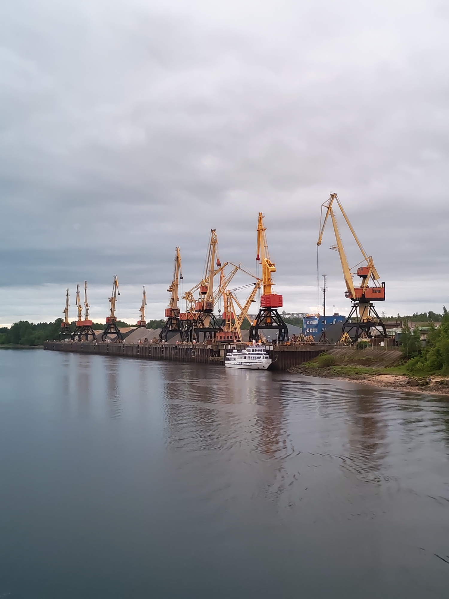 The image depicts a riverfront scene in Bol'shie Mkhi, Russia, under an overcast sky. A series of approximately seven large industrial cranes are stationed along a concrete pier. The cranes have a yellow and orange color scheme, with dark gray support structures. Piles of gray aggregate material are positioned behind the cranes on the pier. A white passenger boat is docked alongside the pier, near one of the cranes. The river is calm, reflecting the sky. A grassy bank borders the river on the right side of the image, with some visible green vegetation. In the background, beyond the pier and cranes, are some industrial buildings and structures.