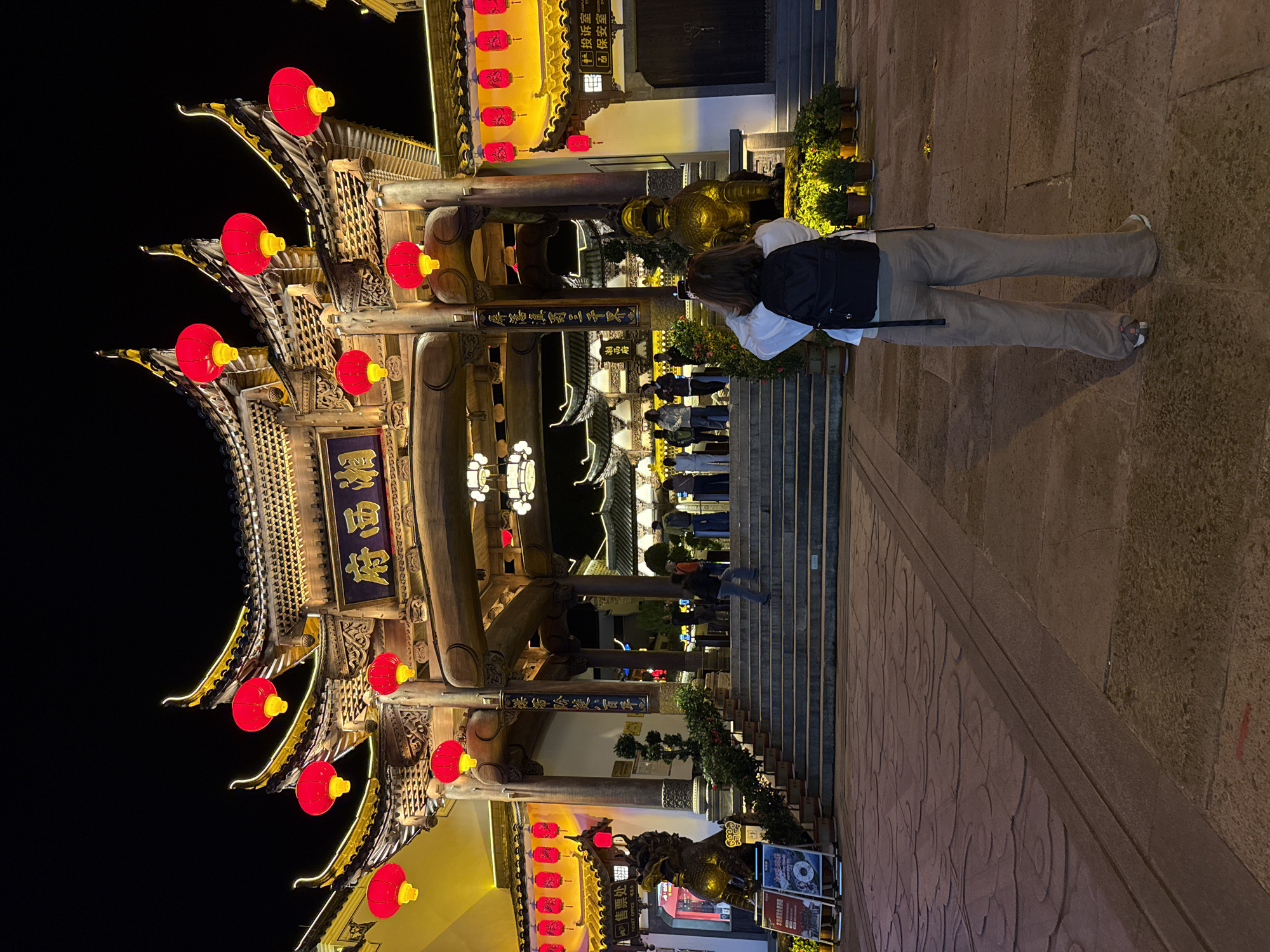 The image depicts a nighttime scene at a traditional Chinese architectural complex, likely a gate or archway, in Wulingyuan, China. A person, viewed from behind, stands in the foreground, facing towards the elaborately decorated structure. This individual wears a white top, light gray trousers, and a black backpack.
The main subject is a multi-tiered traditional Chinese gate, richly adorned with numerous glowing red lanterns. The architecture features intricate carvings and is highlighted with yellow illumination along its eaves and structural elements. Golden statues, possibly of deities or mythical creatures, are visible at the base of the gate's pillars and near the entrance.
To the right of the person, a set of stone steps leads upwards to another part of the complex, where several other people are faintly visible. Potted green plants are placed along the base of the gate and steps. The ground is paved with dark stone or cobblestones. The dark sky confirms the nighttime setting, with artificial lights providing the sole illumination for the scene.
Visible text in Chinese characters includes "售票处" (Ticket Office), accompanied by "CKET OFFICE 매표소" (English and Korean for Ticket Office) on a sign. A prominent blue plaque on the gate displays "湘西府" (Xiangxi Prefecture/Government). Other signs within the complex show "投诉室" (Complaint Room) and "保安室" (Security Room). Vertical plaques on the gate also bear the phrases "■屏藩鎮罰三千界" and "茶香玲進八百平".