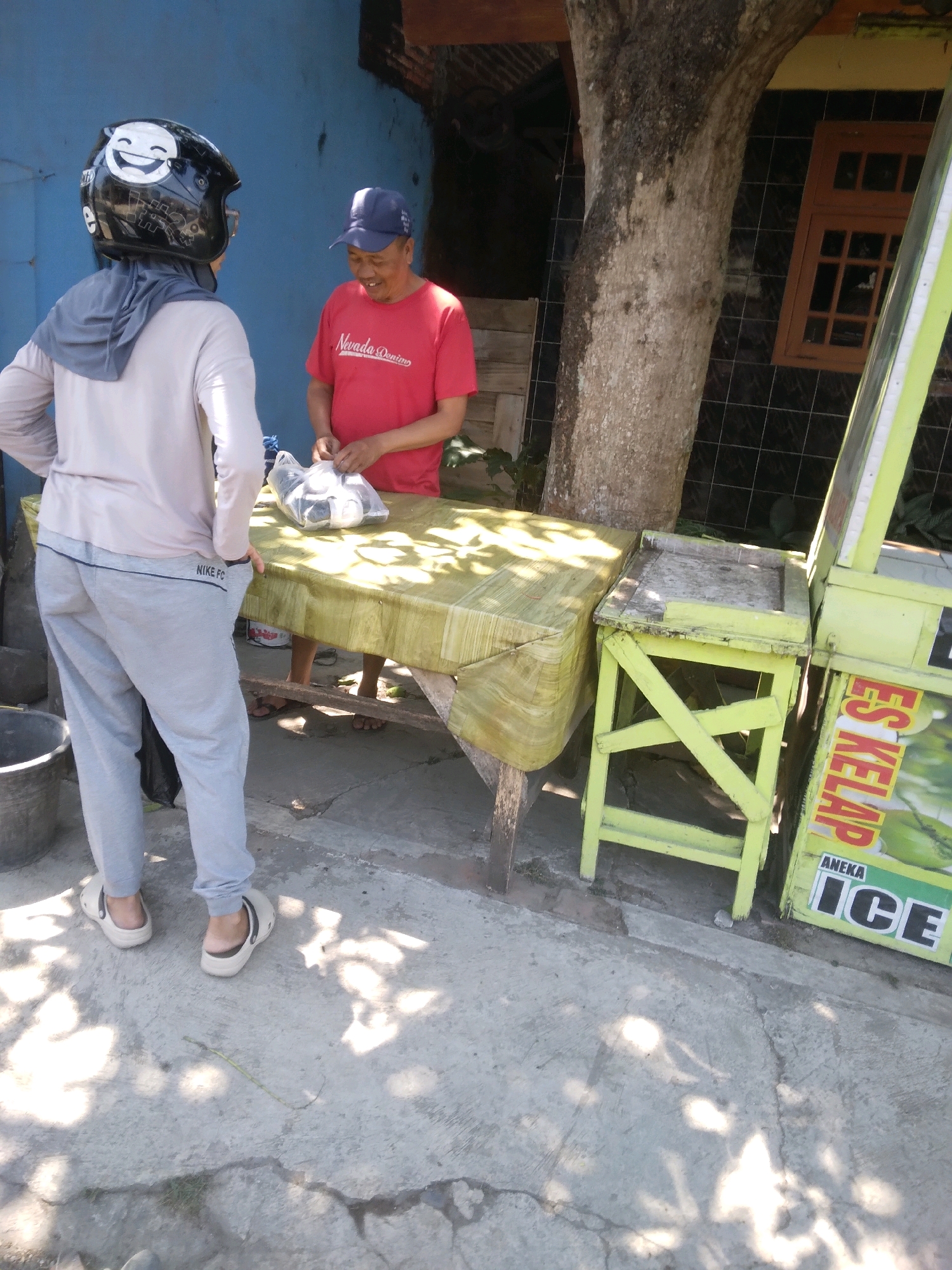 In Sokaraja, Indonesia, a person wearing a grey hijab and a light-colored long-sleeved shirt and grey sweatpants with "NIKE FC" printed on the left leg, stands facing a stall. They are wearing white clogs. A man in a red t-shirt and a blue baseball cap stands behind a table covered with a green cloth, interacting with the person. The man is holding a plastic bag containing items. The stall has a yellow frame and a sign with "ES KELAPA" and "ANEKA ICE" visible. The scene is outdoors, with a large tree trunk to the left of the stall. The ground is paved with concrete, and sunlight casts shadows. A dark grey bucket is to the left of the person. The background shows a blue wall and a wooden structure with a window.