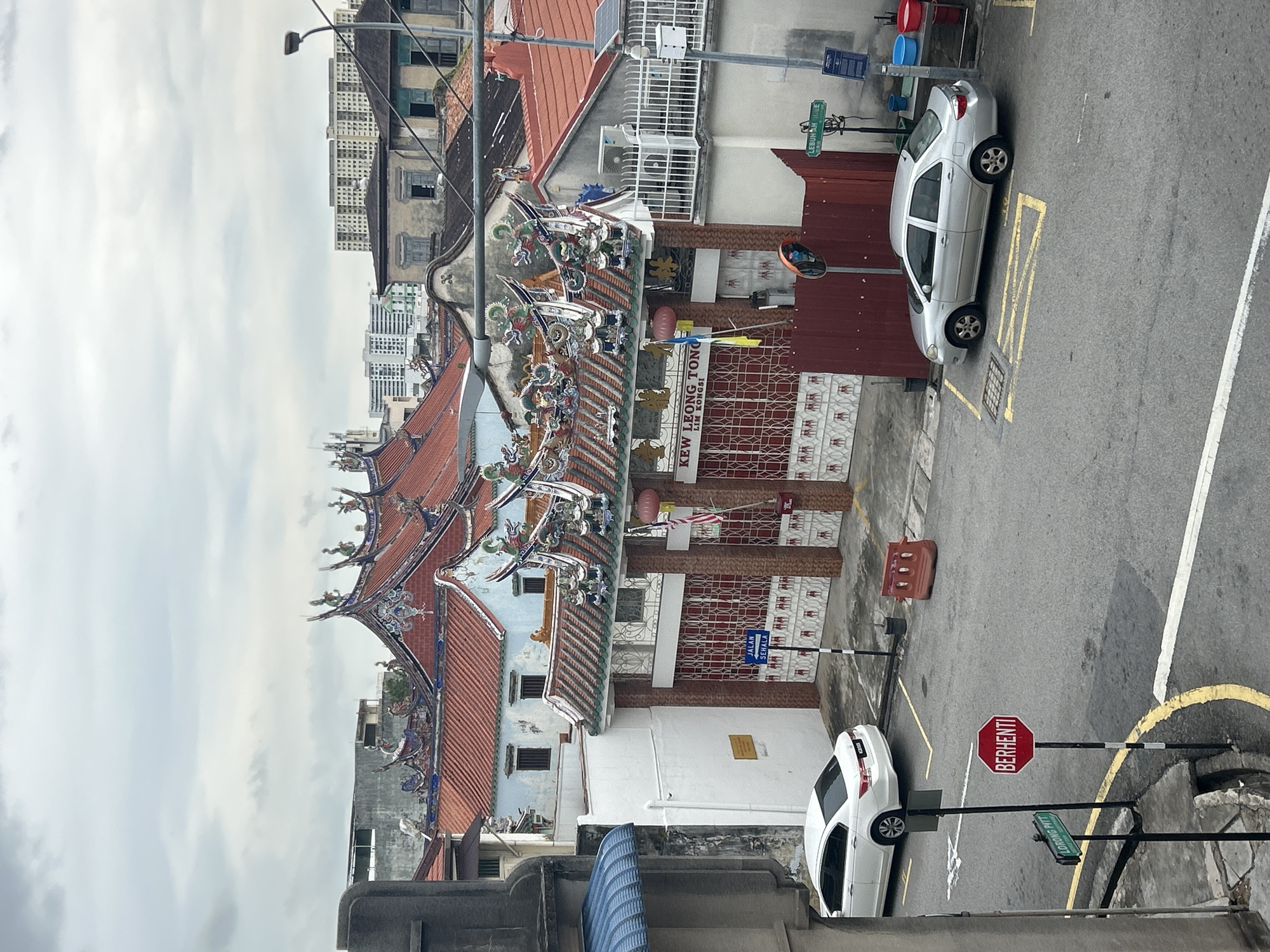The image shows a street scene in George Town, Malaysia, on an overcast day. In the foreground, a silver sedan is parked on the street. Behind it, a white sedan is parked in a lot. To the right, a multi-story building with a red tiled roof and ornate decorative elements stands prominently. The building has a large gate with metal bars. A street sign for "JALAN SEHALA" is visible near the gate, and another sign for "LEBUH AH HUEE" is on the right. In the distance, more buildings, including some taller ones, are visible against the cloudy sky. The street is paved with asphalt and marked with yellow lines. There are several traffic cones and trash bins present.