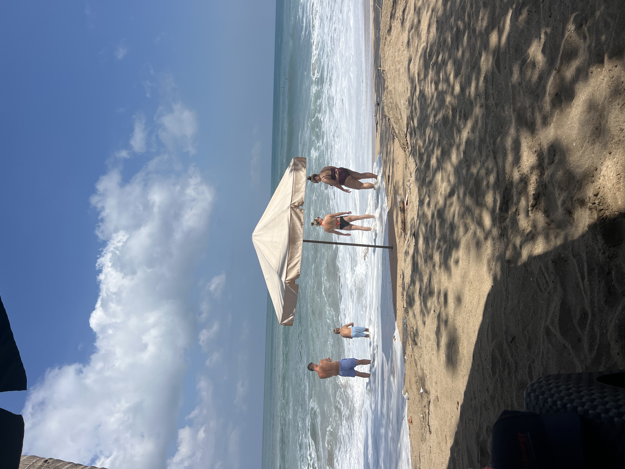 In Kuta, Indonesia, four people are seen standing in the ocean. A man and a child are on the left, facing the water. Two women are on the right, also facing the ocean. An umbrella stands between the two groups, casting a shadow on the sandy beach. The waves are crashing, and there is white foam on the water's surface. The sky is blue with scattered clouds. The scene appears to be a typical day at the beach in Kuta.