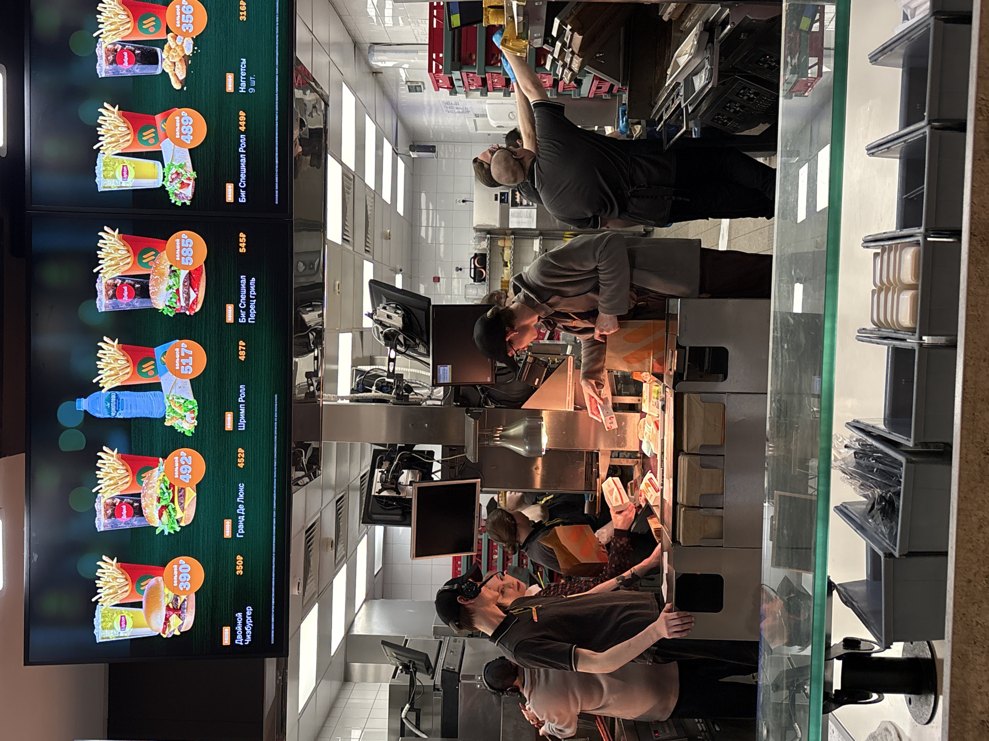 The image shows the interior of a fast-food restaurant in what appears to be Izhevsk, Russia, based on the signage. On the left, there is a digital menu board displaying various meal combinations with prices in Russian Rubles. The menu items include burgers, chicken nuggets, drinks, and wraps. Behind a service counter with a glass partition, multiple employees are working. One worker near the back is reaching up, potentially grabbing supplies from a shelf. Other employees are focused on preparing and packaging food orders.