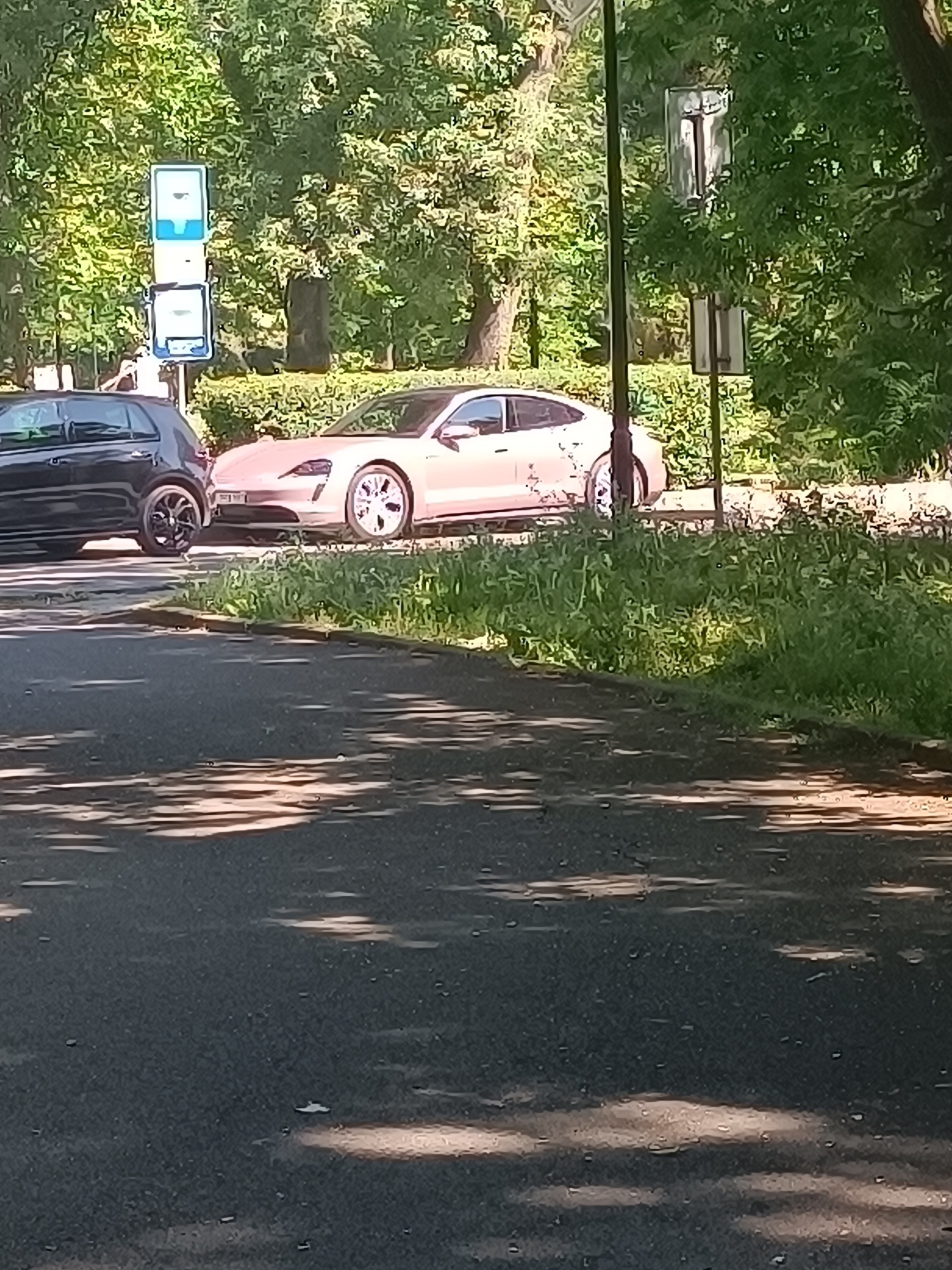 The image depicts a street scene in Burgas, Bulgaria, on a sunny day. Two cars are visible: a black hatchback on the left and a light-colored sedan on the right, possibly a Porsche Taycan based on its shape. Both vehicles are parked or moving slowly along the asphalt road. The background features lush green trees and foliage, indicating a park or a tree-lined street. A blue and white bus stop sign is positioned behind the black car. The road surface shows dappled sunlight and shadow patterns. The overall impression is a tranquil urban setting with vehicular traffic and natural elements.