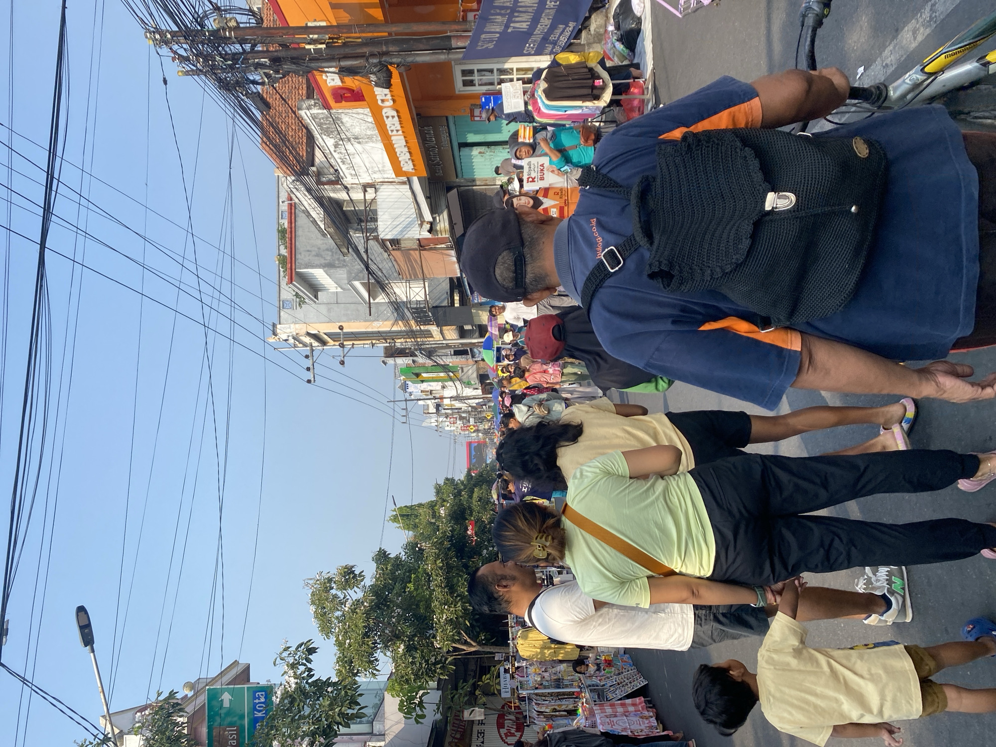 This is an eye-level, medium shot of a bustling street scene in Kediri City, Indonesia, likely during daytime given the bright sunlight and clear blue sky. The street is lined with buildings on both sides and is filled with a crowd of people, suggesting a market or a public event. The perspective is from behind a person wearing a dark blue t-shirt and a black crocheted backpack, who is holding onto the handlebars of a bicycle.
In the foreground, to the left, a young boy in a light yellow t-shirt and brown shorts is being led by the hand by an adult, likely his parent. Behind him, a man in a white t-shirt and grey shorts is walking, and to his right, a woman in a light green t-shirt and black pants is walking with her back to the camera. Further down the street, more people are visible, some browsing through stalls set up along the roadside displaying various goods, including clothing on racks.
The buildings lining the street have a mix of architectural styles, with some featuring orange and white facades. Overhead, a tangle of electrical wires and poles are a prominent feature of the urban landscape. A street sign with green and white text, partially obscured by trees, is visible on the left side of the image. The ground is paved, indicating a city street. The overall atmosphere is one of lively activity and a community gathering.
