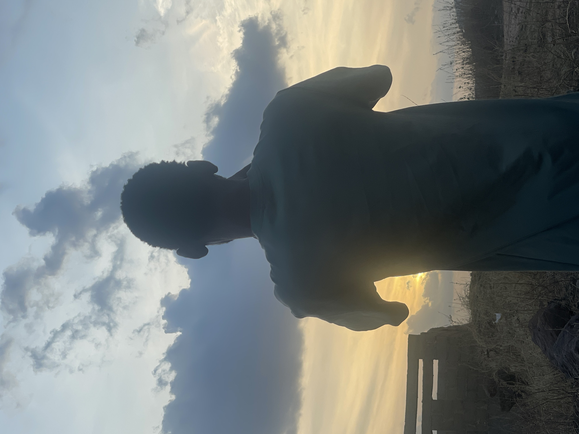 This image captures a silhouette of a person against a dramatic sky, likely at either sunrise or sunset in Lokoja, Nigeria. The photograph is taken from a low angle, emphasizing the vastness of the sky and the sun's warm glow.
The main subject is a person, seen from the back and left side, standing upright and looking towards the right side of the frame where the sun is setting or rising. Their form is a dark silhouette against the bright backdrop, making details of their face and clothing indistinguishable, though they appear to be wearing a long-sleeved garment.
The sky dominates the upper portion of the image, showcasing a striking transition of colors. On the left, the sky is a soft, light blue-grey with several dark, textured clouds. Towards the right, it shifts into a radiant yellow and orange hue, indicating the direct presence of the sun breaking through a lighter cloud layer. This bright glow strongly suggests either early morning or late evening.
In the lower right background, sparse, dry vegetation and what appears to be a dark, block-like structure or rock formation are visible, also in silhouette. This landscape suggests a rural or semi-arid environment. The overall mood is one of quiet contemplation or appreciation for the natural beauty of the sky. There is no visible text in the image.