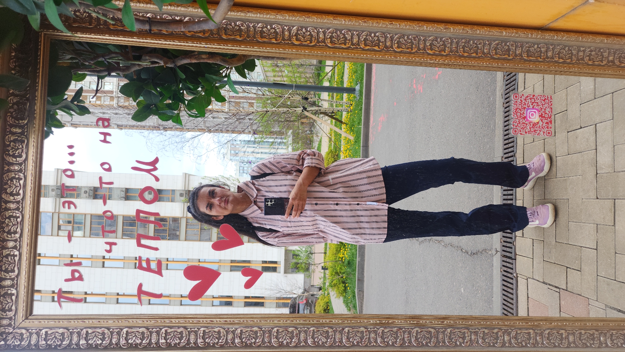 A young woman is reflected in a large, ornate, rectangular gold-colored mirror in Moscow, Russia. She is wearing a horizontally striped pink and white shirt, black pants, and pink sneakers. She holds a black smartphone in her left hand. The mirror also reflects a cityscape, including a tall building with many windows, greenery, a paved area, and a QR code associated with Instagram. In the reflection on the mirror are red hearts and Cyrillic writing. The frame around the mirror is heavily decorated with an intricate pattern. The location appears to be outdoors, possibly a park or urban area.