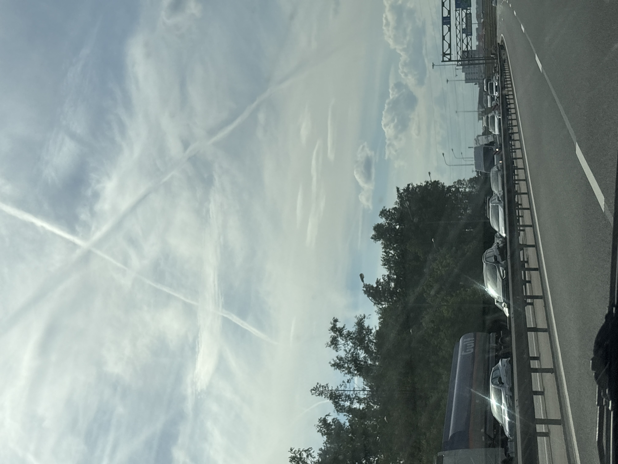 The image depicts a daytime highway scene in Novosergievka, Russia, characterized by vehicle traffic and a sky marked with aircraft contrails. The perspective is from within a vehicle, showing the road ahead.
The upper two-thirds of the frame are dominated by a bright, light blue sky with scattered white clouds, primarily cirrus and altocumulus formations. Multiple long, thin white aircraft contrails are visible, with two intersecting prominently in the upper-center to form an 'X' shape. Additional contrails extend diagonally across the sky.
The highway itself is a multi-lane asphalt road with visible white lane markings. A metallic safety barrier runs along the left side of the highway, separating the viewer's lane from adjacent traffic. To the left of this barrier, a lane of vehicles is in heavy congestion or stopped. A prominent dark blue tanker truck, bearing the word "Gulf" on its side, is visible on this side of the road. Behind it, several passenger cars, including a silver sedan and a dark SUV, reflect sunlight from their surfaces.
An embankment covered with dense green trees and foliage borders the highway on the left. Several streetlights extend above the treeline. In the distance, on the right side of the highway, more vehicles, including a white van and multiple passenger cars, are observed in traffic. Overhead gantries with unreadable road signs are present further down the road on the right, accompanied by indistinct structures suggesting urban or industrial infrastructure in the background. No individuals are explicitly visible.