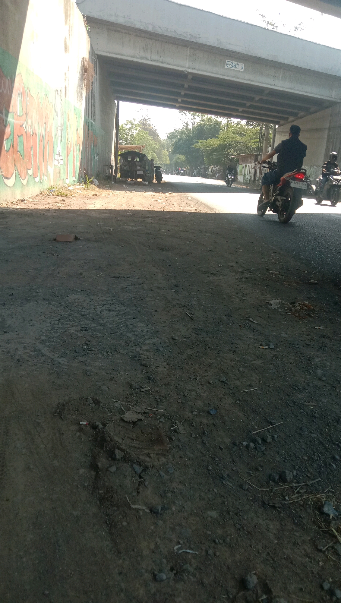 The image depicts a street scene under an elevated concrete structure, likely an overpass, in Karang Anyar, Indonesia. The foreground is a rough, dirt-and-gravel road surface with tire tracks visible. To the left, a concrete wall is covered in colorful graffiti. Beyond the wall, a small, open-bed vehicle, possibly a modified car or a small truck, is parked. Several motorcycles are also present. In the middle ground, a man on a motorcycle is riding away from the camera, towards the underpass. Another motorcycle and rider are visible further down the road, also moving away. Trees and buildings line the road in the distance. The lighting suggests it is daytime, with sunlight casting shadows. A discarded cardboard box is on the ground in the foreground.