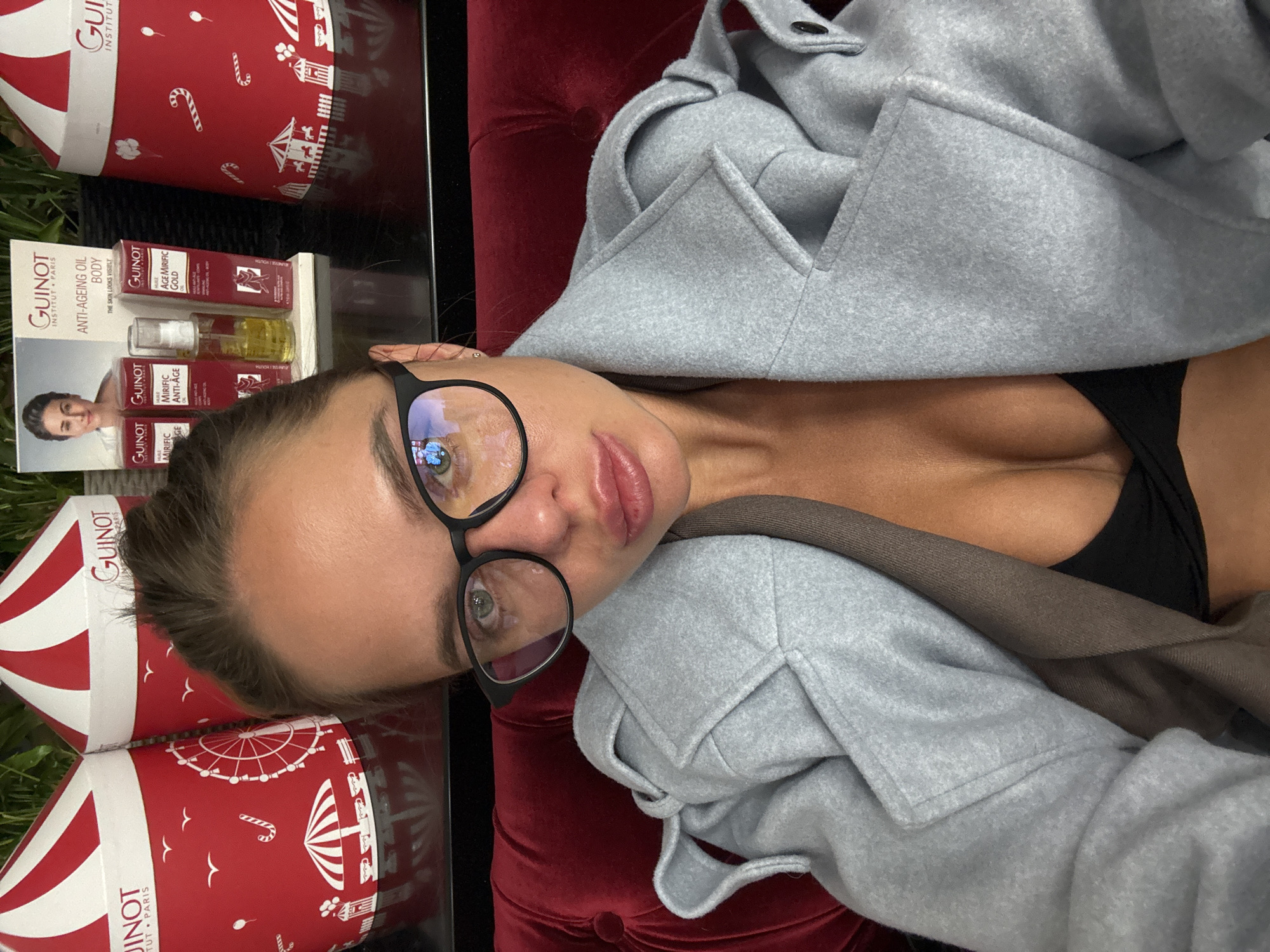This media file, taken in Moscow, Russia, presents a close-up, rotated portrait of a woman. She is shown reclining or lying down, with her face and upper torso partially visible on the right side of the frame. She is wearing black-framed glasses, a light grey coat, and a black top or bra. Her skin has a tanned tone, and her lips are slightly parted with a neutral expression.
Prominently featured in the background and foreground are several red and white cylindrical containers, likely gift boxes or product packaging. These containers are branded with "GUINOT INSTITUT PARIS" and decorated with festive, carnival-themed imagery such as carousels, Ferris wheels, and candy canes. One product, specifically identified as "GUINOT ANTI-AGEING OIL BODY," is visible in its packaging, which includes two small bottles of oil. Additional text on visible products includes "MIRIFIC AGE GOLD," "MIRIFIC ANTI-AGE," and the French word "HUILE" (oil).
The woman appears to be resting on a plush red velvet surface, possibly a chair or sofa, which forms part of the background. Reflections in her glasses show indoor lighting. The overall context suggests a focus on beauty, skincare products, or a promotional setting for the Guinot brand. The exact time of day and weather are not discernible due to the indoor setting.