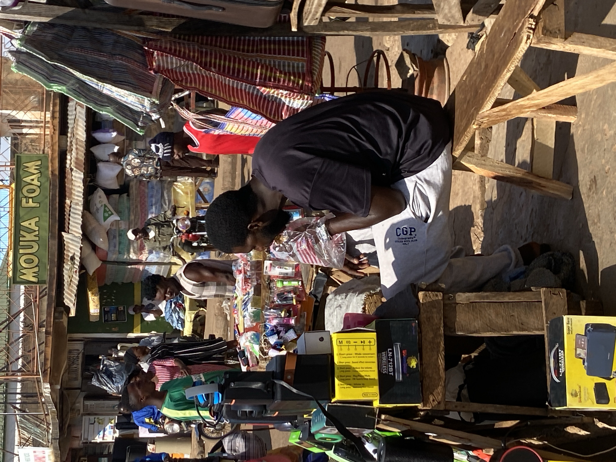 The image depicts a street scene in Bukuru, Nigeria. A man wearing a black shirt and gray sweatpants bends over a display of small items, possibly for sale. The display includes various colorful objects, electronics packaging, and other goods. Behind him, a wooden structure and assorted textiles are visible. Other people can be seen in the background, along with storefronts and signs, one of which reads "MOUKA FOAM." Motorbikes are also present. The scene suggests a busy, open-air marketplace.