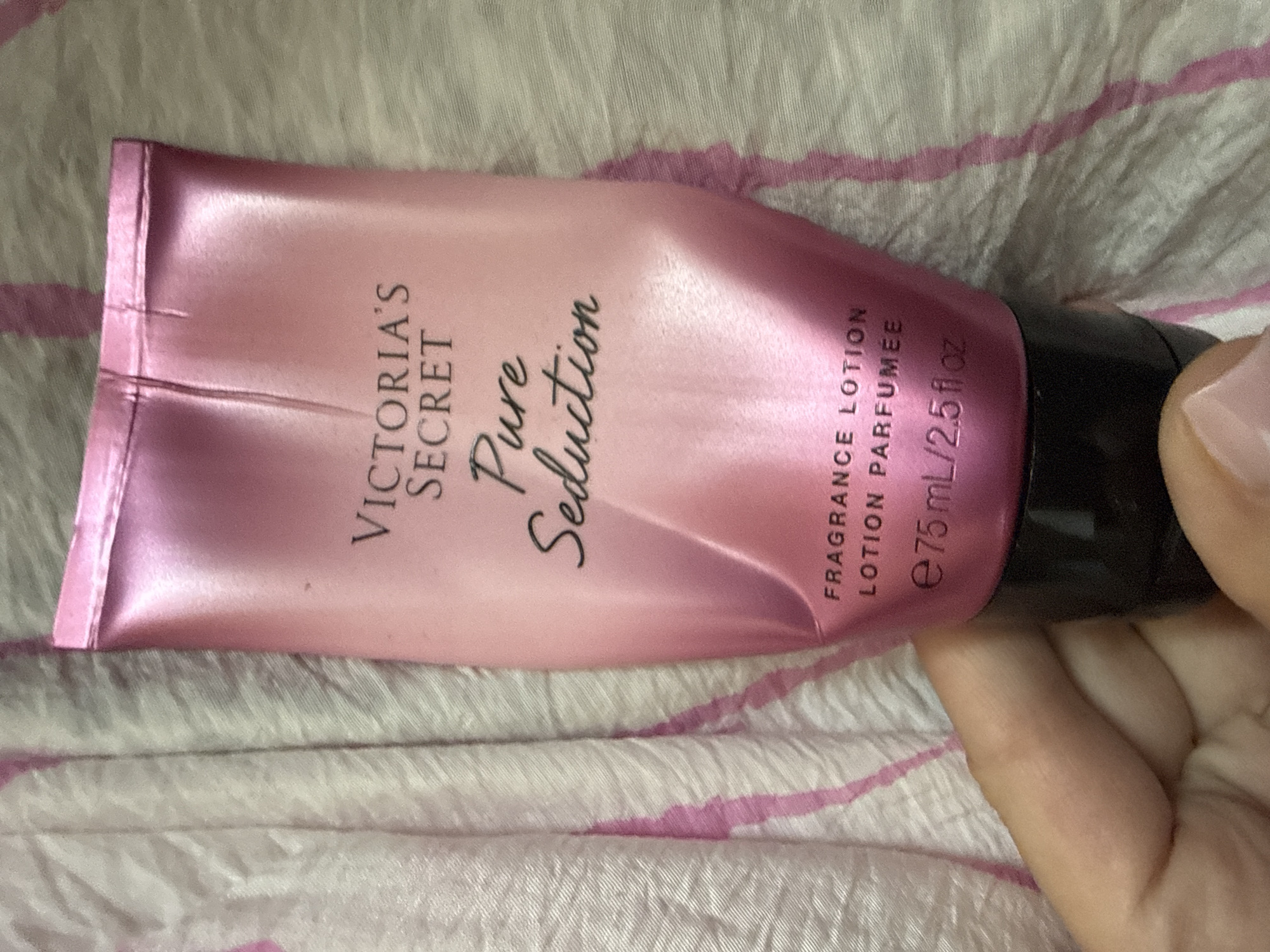 A person's hand is holding a pink tube of Victoria's Secret Pure Seduction fragrance lotion. The tube is metallic pink and has black text on it. The background is a pink and white striped fabric. The image does not contain any information about the city of Okhtyrka, Ukraine.