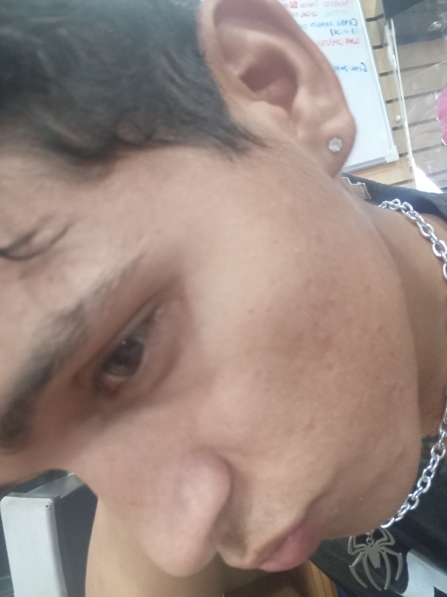 A close-up, slightly angled, indoor photograph depicts a person with brown skin and dark hair. The person appears to have a single earring in their left ear. A silver chain necklace with a spider pendant is visible around their neck. The background shows a blurred white board with writing and various indistinct objects. No information suggests a location in Juan Griego, Venezuela.
