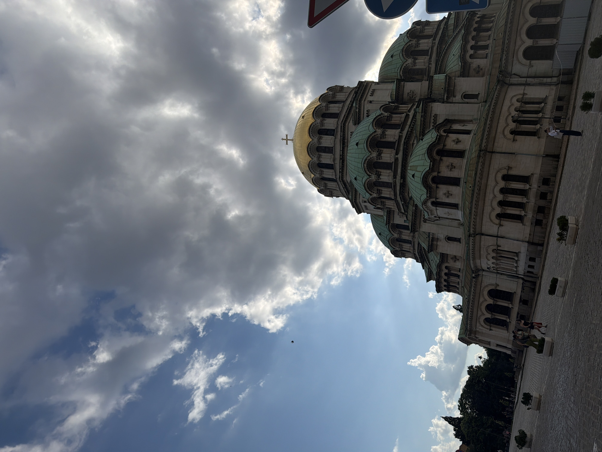 The image depicts the St. Alexander Nevsky Cathedral in Sofia, Bulgaria, captured from a low, slightly tilted perspective. The grand cathedral dominates the right half of the frame, featuring a prominent golden central dome topped with a cross, surrounded by numerous smaller, weathered green-patina copper domes and arched windows on its light-colored stone facade.
The sky is a dramatic mix of conditions. The upper right and central portions are covered by large, dark grey, turbulent clouds, through which shafts of sunlight occasionally break. The upper left quadrant reveals a clear blue sky with scattered white cumulus clouds, indicating a partly cloudy day. A small, dark bird is visible in flight against the blue sky in the upper left.
The ground consists of a cobblestone plaza, partially visible in the lower half of the image. Several rectangular white planters with small green shrubs are spaced across the plaza. On the left side, dark green trees are visible in the distance.
Several individuals are present on the plaza: a lone person in a white shirt and blue pants stands near the base of the cathedral on the lower right, and a group of two or three people walk further back towards the left. A red-bordered triangular traffic sign and a blue circular sign are partially visible on the far right edge of the frame. A construction barrier or tape is discernible near the base of the cathedral where the lone person stands.