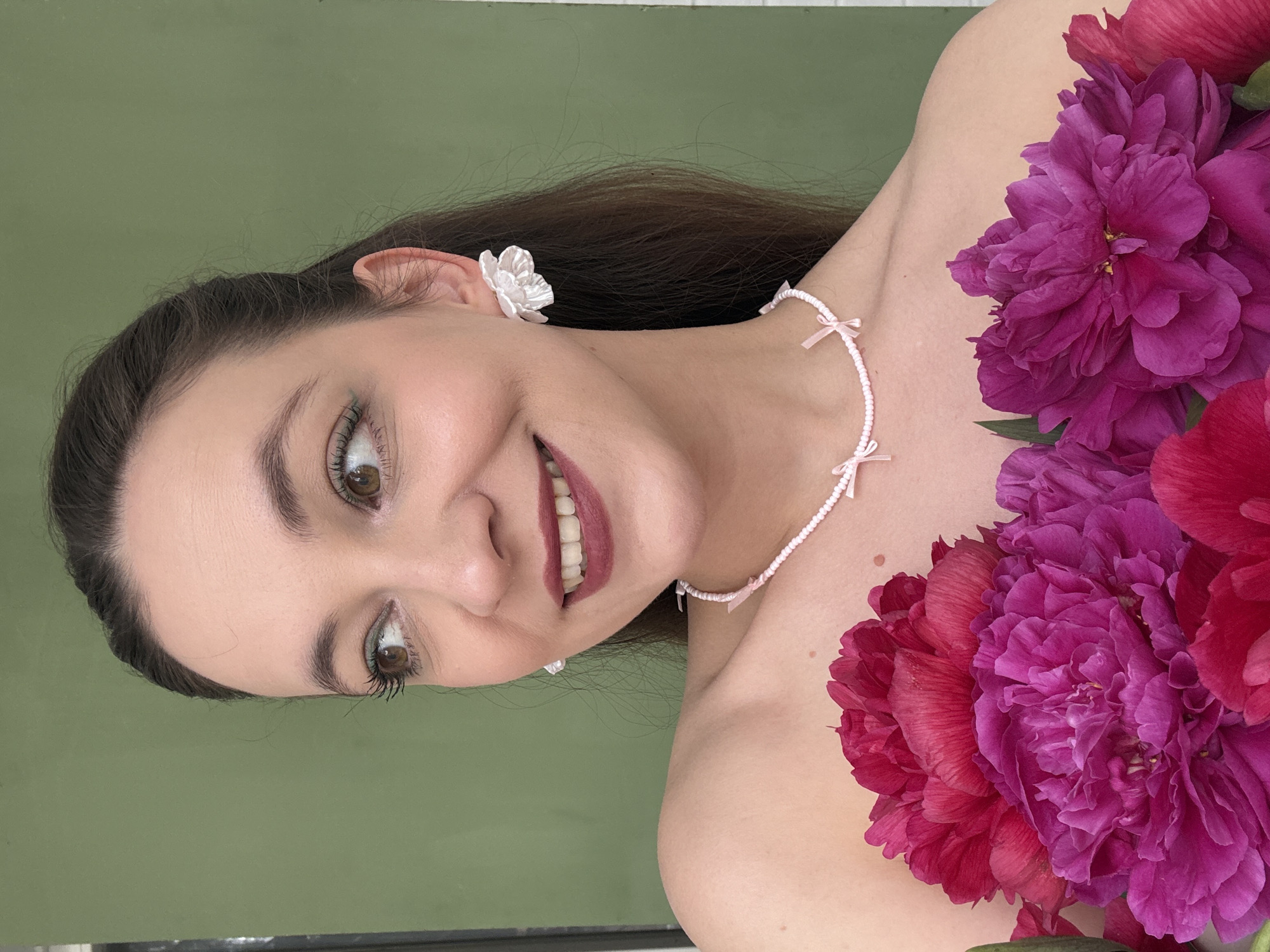 In this close-up, vertically oriented media file, a woman with dark hair and brown eyes is the central subject. She is adorned with delicate floral earrings and a pearl necklace with pink bows. She is also partially obscured by lush pink and magenta peonies, which appear to be draped across her shoulder and chest. The woman is smiling, displaying white teeth, and her eyes are looking to the left of the frame. Her makeup includes dark mascara and dark pink lipstick.
The background is a solid, muted green wall. The lighting suggests it is daytime, possibly indoors given the controlled lighting and the absence of harsh shadows. The overall mood conveyed is one of gentle beauty and perhaps a celebratory or romantic aesthetic, emphasized by the flowers and feminine jewelry. No text is visible in the image. The location context is Chaikovsky, Russia, though this is not visually evident from the image itself.