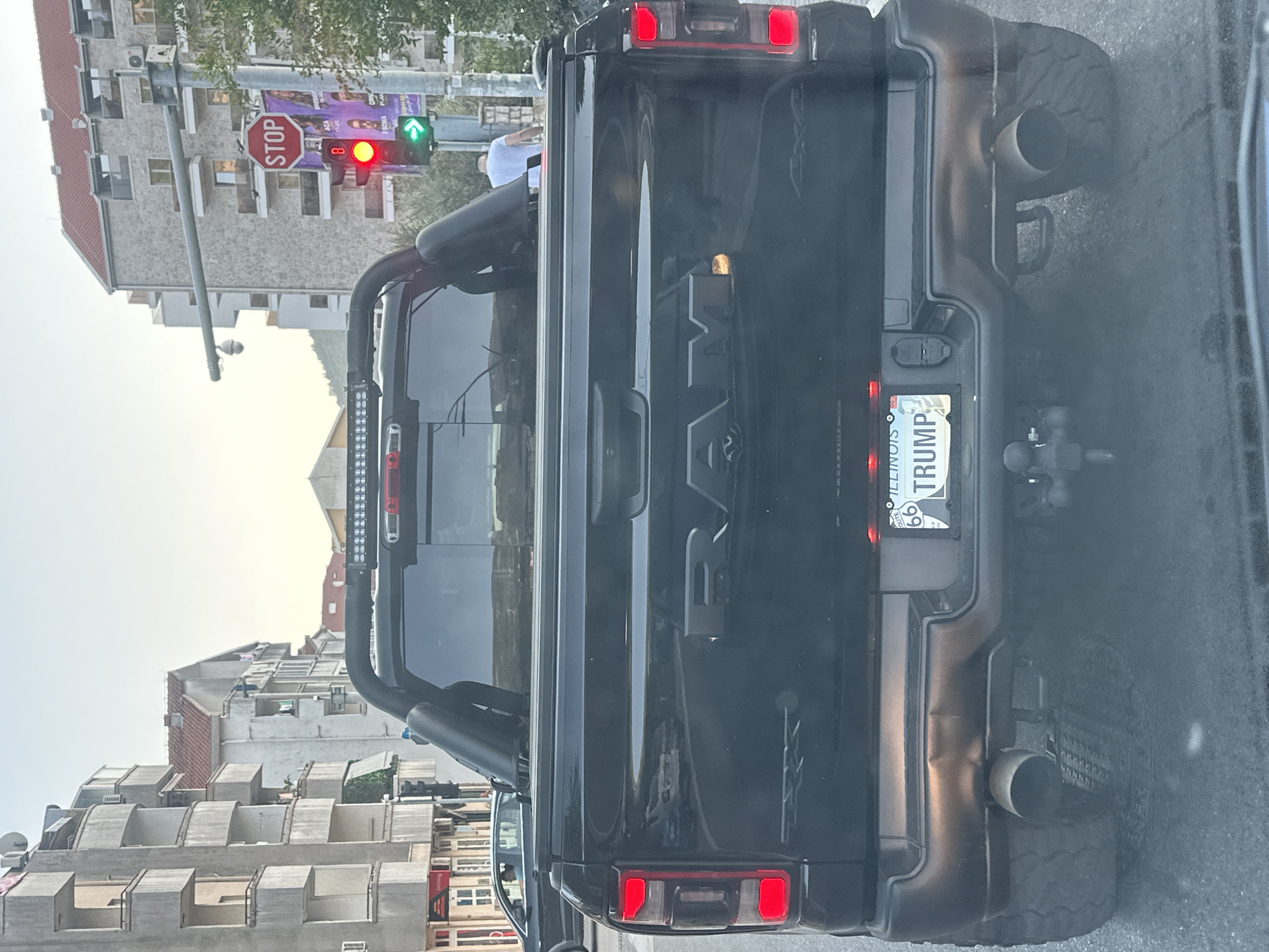 The image shows the rear of a black Ram pickup truck. The truck has a license plate that reads "ILLINOIS" and "TRUMP". The truck is in traffic on a city street, and a traffic light is visible in the background. Buildings and a stop sign are also visible in the background, consistent with a city setting in Budva, Montenegro. The overall scene is a street view with a focus on the vehicle's rear.