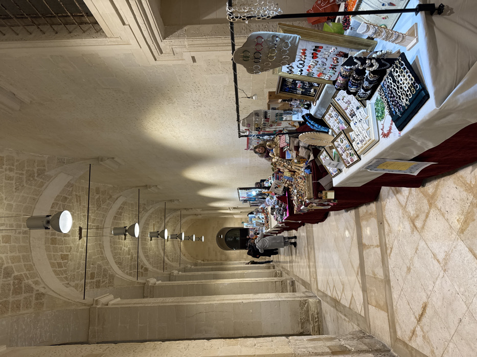 This image depicts a long, vaulted corridor within a historic building in Lecce, Italy, functioning as an indoor market. The corridor features robust, light-colored stone pillars and arches supporting a similarly constructed vaulted ceiling, from which a series of modern industrial-style pendant lights are suspended. The floor is tiled with large, light-colored stone or marble in a geometric pattern.
Along the right side of the corridor, a continuous line of temporary market stalls is set up, extending into the distance. Each stall consists of tables draped with dark red and white tablecloths, laden with a variety of handcrafted items, predominantly jewelry such as necklaces, earrings, bracelets, and rings displayed on various stands, trays, and busts. Small decorative objects are also visible.
Several individuals are present within the corridor. In the mid-ground on the right, a woman with dark hair, wearing a dark top, is positioned behind a market stall, smiling directly towards the viewer. Further down the corridor, a person wearing a long light-grey coat is walking away from the camera. Additional indistinct figures are visible deeper within the corridor, appearing to observe the stalls or move through the space. A high, barred window opening is visible in the stone wall on the right side in the background. The scene is well-lit by the overhead fixtures, casting distinct light patterns on the walls and ceiling.