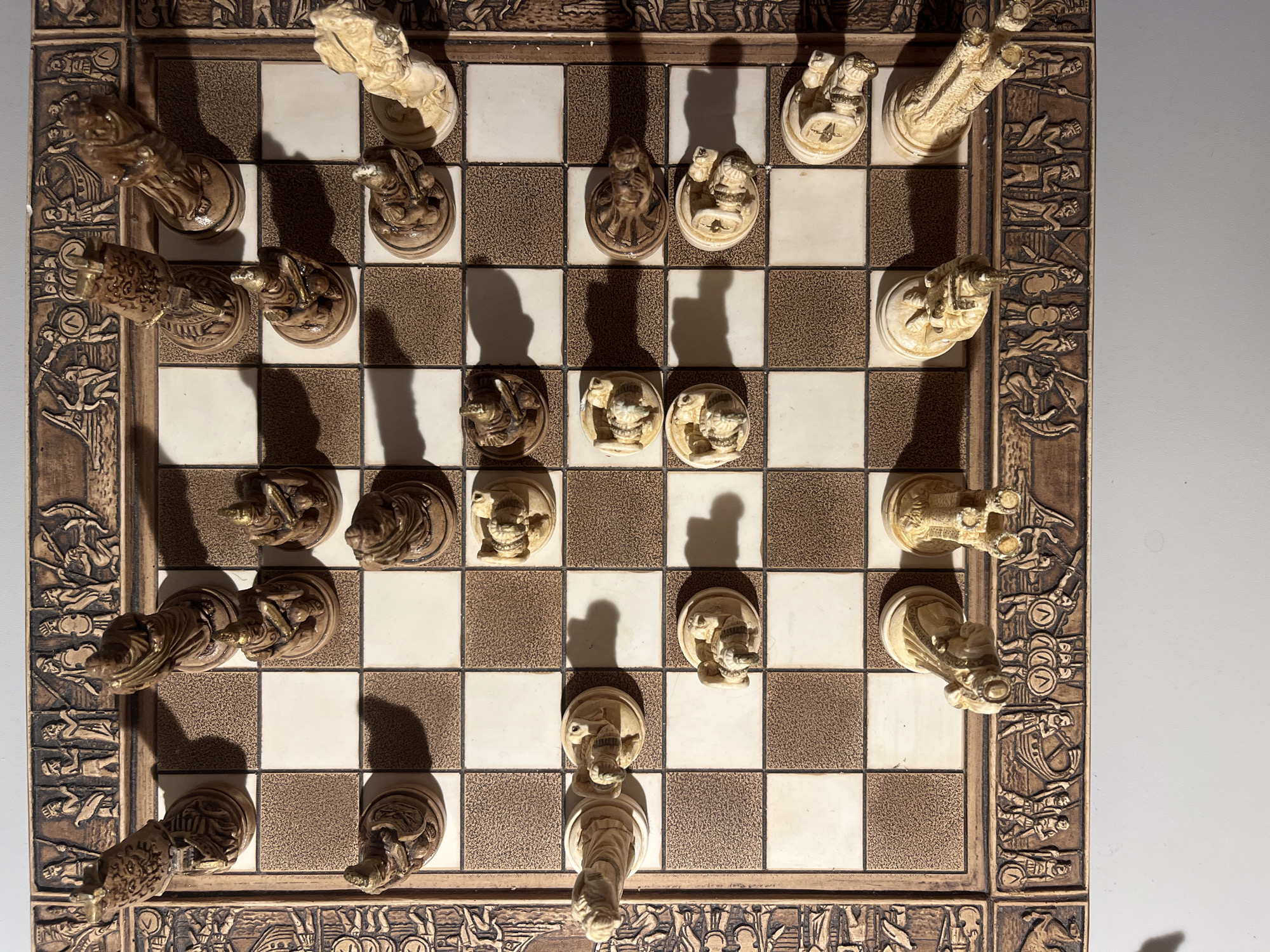 The image shows a chess board with pieces, set on a table. The chess pieces appear to be made of carved wood or resin and are in various states of play. The board itself is bordered by decorative carvings. The scene is well-lit, with shadows cast by the pieces. There are no people visible in the image. The location is not specified and cannot be determined.