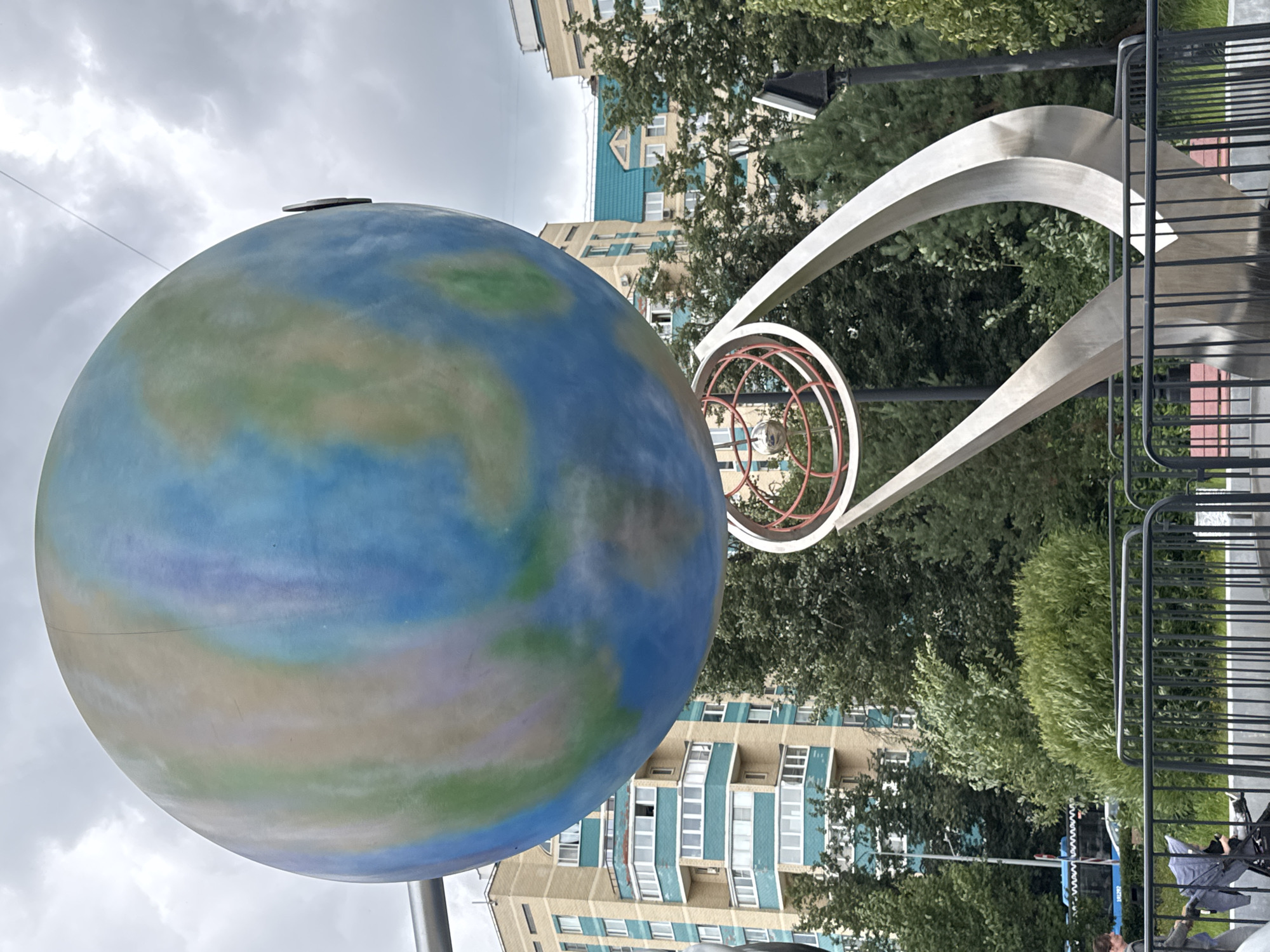 A large, stylized globe sculpture painted in shades of blue, green, and beige stands prominently in the foreground against a cloudy sky. Below the globe, a metallic structure with red concentric rings and a spherical element is visible. In the background, multi-story residential buildings with beige and teal facades are situated behind a dense arrangement of green trees. A black metal fence runs across the lower part of the image, with a person pushing a stroller visible on the left side. A blue bus with white lettering is partially visible to the left of the trees. The scene appears to be an outdoor public space in the city of Zelenograd, Russia.