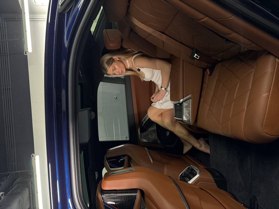 The image depicts a female subject seated in the rear passenger compartment of a luxury vehicle, located in Warsaw, Poland. The woman, appearing to be in her twenties, possesses long blonde hair and is dressed in a white sleeveless slip dress, a white choker necklace, and a silver watch on her left wrist. She wears light-colored high-heeled shoes. She is positioned on the right-hand side rear seat, with her left leg bent and her right leg extended and crossed over the left. Her hands are clasped in her lap, and she gazes slightly to her left.
The vehicle's interior features rich brown leather upholstery with diamond-quilted stitching and perforated accents on the seats. A dark blue metallic finish is visible along the car's upper exterior frame. The front passenger seatback is equipped with an integrated display screen, presenting a dark, possibly carbon fiber, texture and a distinct red and blue striped emblem. A central rear armrest, partially deployed, includes metallic-trimmed controls. An overhead sunroof is visible. The external environment, seen through the car's windows and open door frame, consists of an indoor setting characterized by dark gray walls, exposed ceiling structures, and overhead white fluorescent strip lighting, indicative of a professional showroom, studio, or underground parking facility.