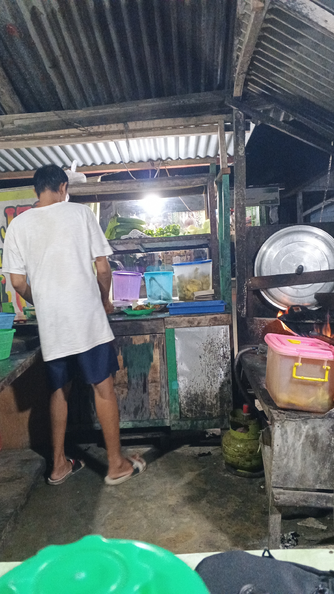 The image depicts an outdoor food stall at night in Tumang, Indonesia. A person wearing a white t-shirt and dark shorts stands facing away from the camera, appearing to be engaged in preparing food at the stall. The stall itself is constructed with rustic materials, including corrugated metal roofing and weathered wooden planks. Behind the person, a counter displays several plastic containers filled with food items and vegetables. To the right, a large pot is being heated over an open flame, with a gas cylinder visible nearby. The lighting is dim, suggesting it is nighttime, and the overall scene conveys a humble, operational food vendor setting.