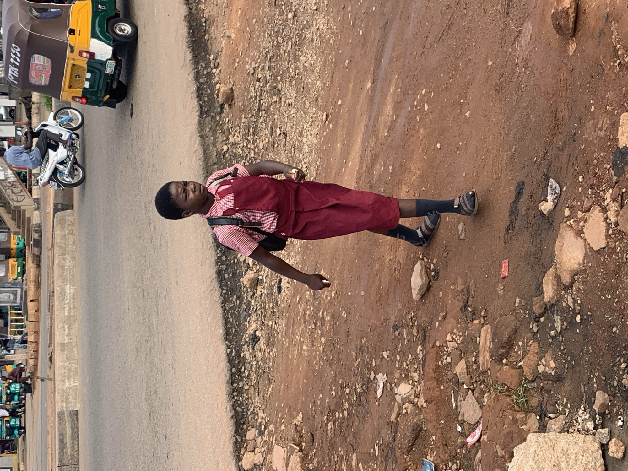 A young Black person in a school uniform, consisting of a red and white checkered shirt and a maroon pinafore, walks along a dirt road in Gura Top, Nigeria. The person carries a black backpack. In the background, a white motorcycle with a rider is visible on a paved road, alongside a yellow and green motorized tricycle. The environment appears to be a roadside, with loose stones and debris scattered on the ground. The lighting suggests it is daytime, with overcast skies.