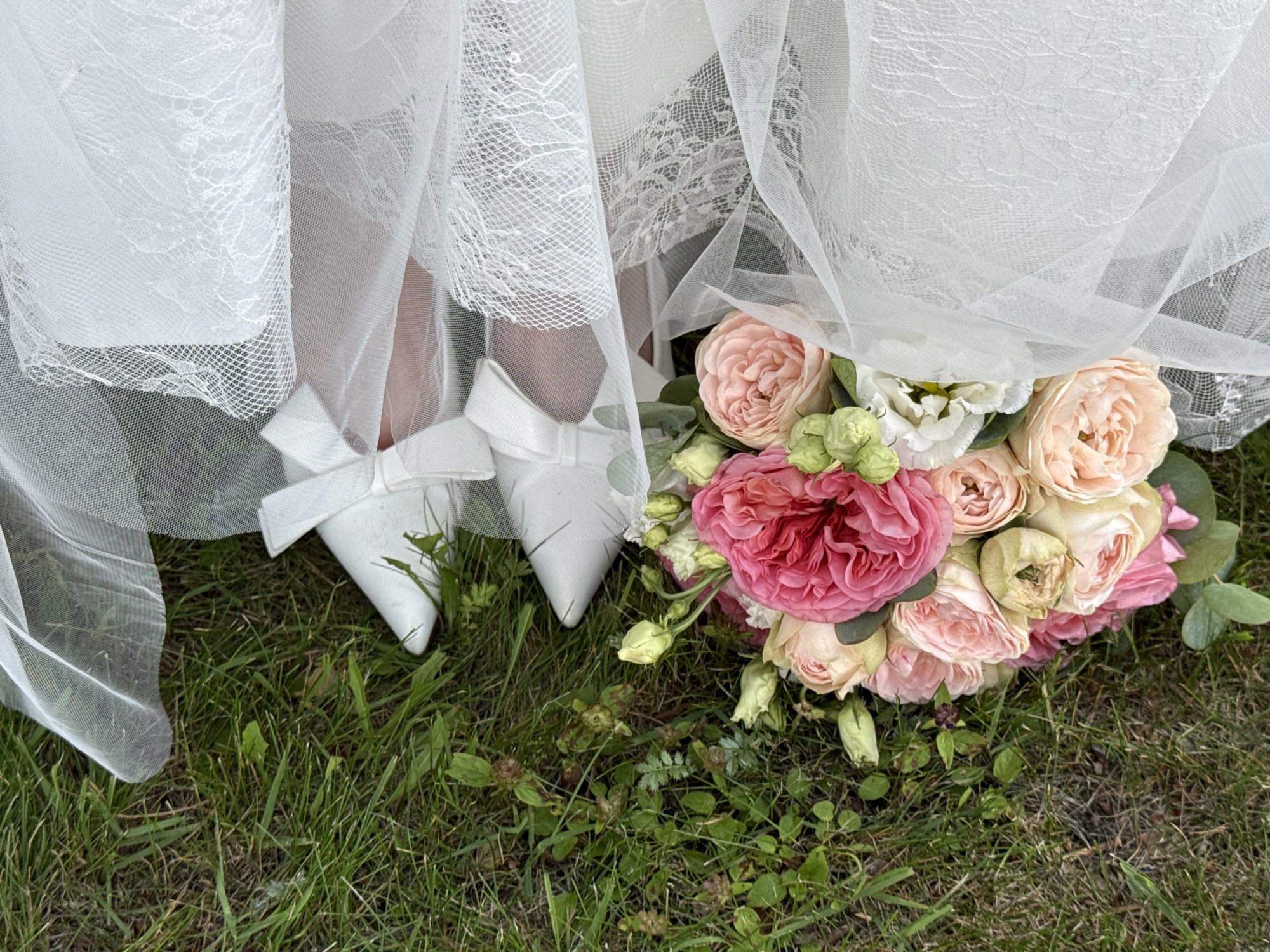 In Votkinsk, Russia, a close-up aerial shot reveals a wedding bouquet resting on green grass. The bouquet consists of various shades of pink and cream roses, with small green buds and foliage. Partially obscuring the bouquet and the grass are layers of white tulle and lace, suggestive of a wedding veil. Also visible are a pair of white satin heels with bows, positioned atop the tulle. The scene is set outdoors, with the focus on the bridal elements arranged on the ground.