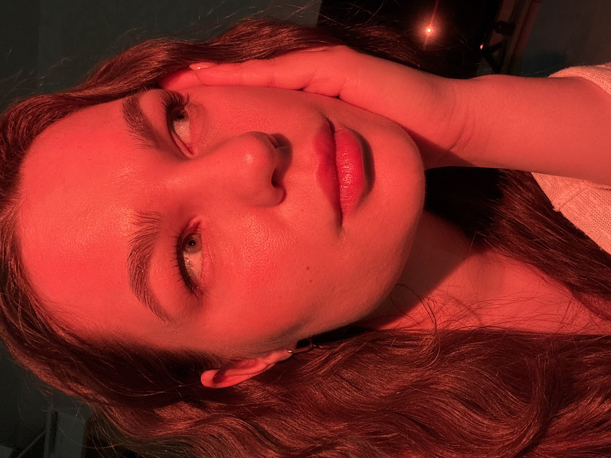 A close-up, rotated portrait of a young woman under strong reddish-orange lighting. Her head is tilted, with her right hand partially obscuring the top of her forehead. Her eyes are open, looking upwards. Her lips are colored with a dark shade of lipstick. Her hair is wavy and dark brown. A small silver earring is visible in her left ear. The background is dark, with a faint light source visible at the top. It's not possible to determine the location in Chaikovsky, Russia from this image alone.