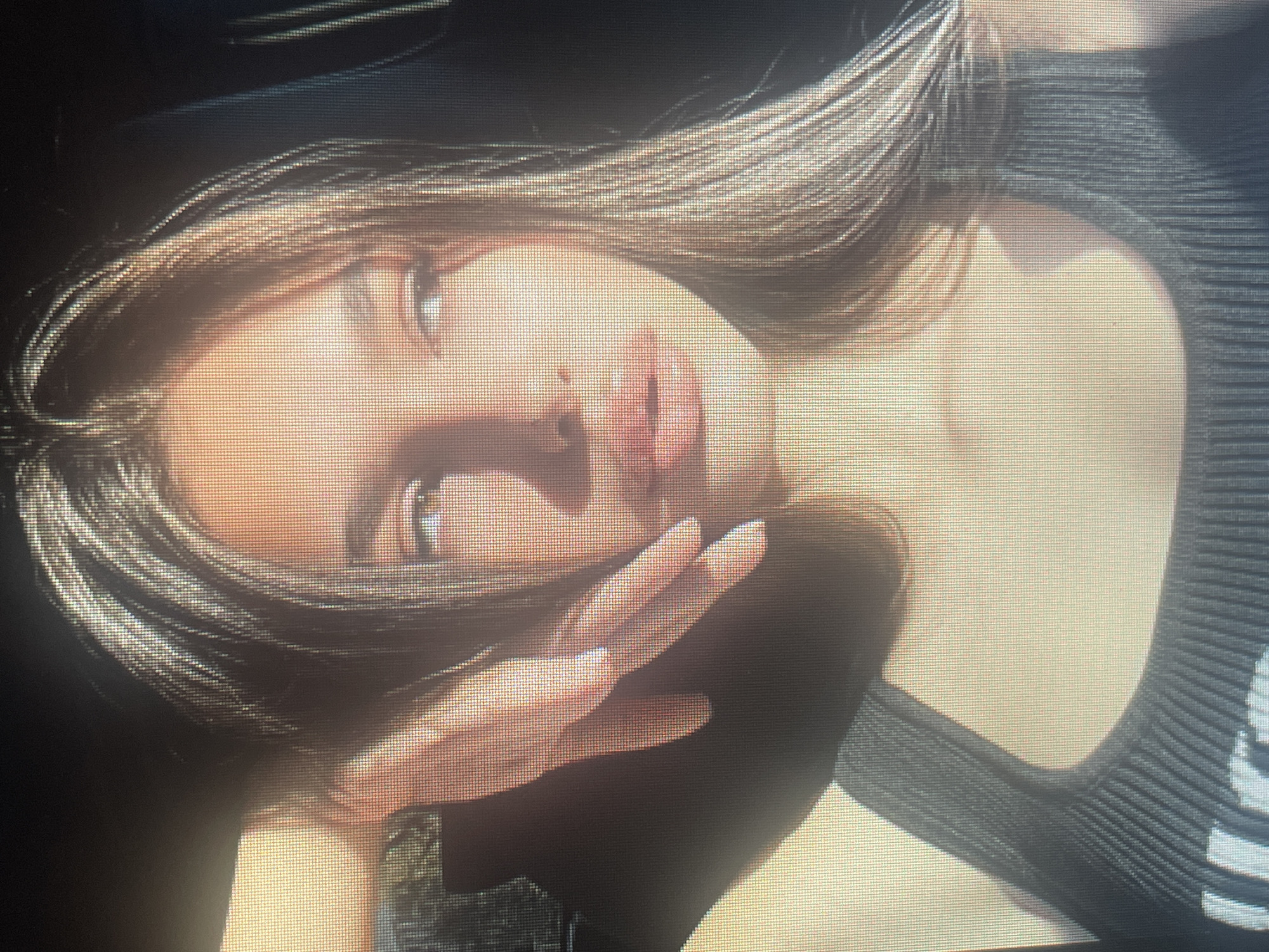 A close-up, eye-level shot captures a young woman from the chest up, with her head turned slightly to her right, looking off into the distance. Sunlight illuminates the right side of her face, casting shadows and highlighting her features. Her hair, a mix of dark brown and lighter streaks, falls past her shoulders. Her left hand is raised, with fingers gently touching her forehead and hair, adding a touch of casual grace. She wears a dark, ribbed tank top. The background is dark and out of focus, suggesting an indoor setting, possibly a car interior due to the angle and the way light enters the frame. The image appears to be taken on a screen, as indicated by the visible pixel grid. The overall mood is serene and contemplative, bathed in the warm glow of natural light. The location of Mantov, Czechia, is not discernible from the image itself, as there are no visual cues to suggest a specific geographic setting within that city.
