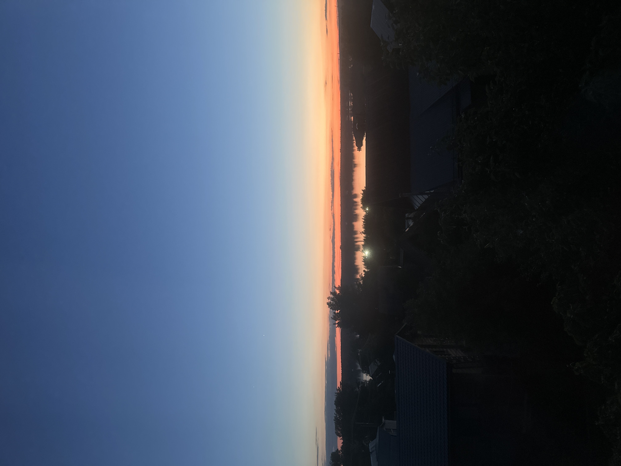 The image presents a serene twilight landscape, likely a sunset, over a wide body of water in Chaikovsky, Russia. The sky, which occupies the upper half of the frame, displays a striking gradient of colors, transitioning from a deep blue overhead to lighter blues, pale yellow, and vibrant orange-pink hues near the horizon, indicating the recent setting of the sun. Two small, distinct bright dots, possibly stars or planets, are visible in the darker blue expanse of the sky.
A calm, reflective body of water stretches across the mid-ground, mirroring the warm tones of the sky. Along the distant shoreline, a dark, silhouetted band of trees and a few structures are visible, punctuated by several bright lights that cast shimmering reflections onto the water's surface.
In the foreground, on the lower right side, dark silhouettes of dense tree foliage and parts of buildings, including rooftops and walls, are visible, suggesting the photograph was taken from an elevated perspective, such as a balcony or window. The scene is devoid of explicit human activity or visible text. The atmosphere is one of tranquility and calm, characteristic of the transition between day and night under clear weather conditions.