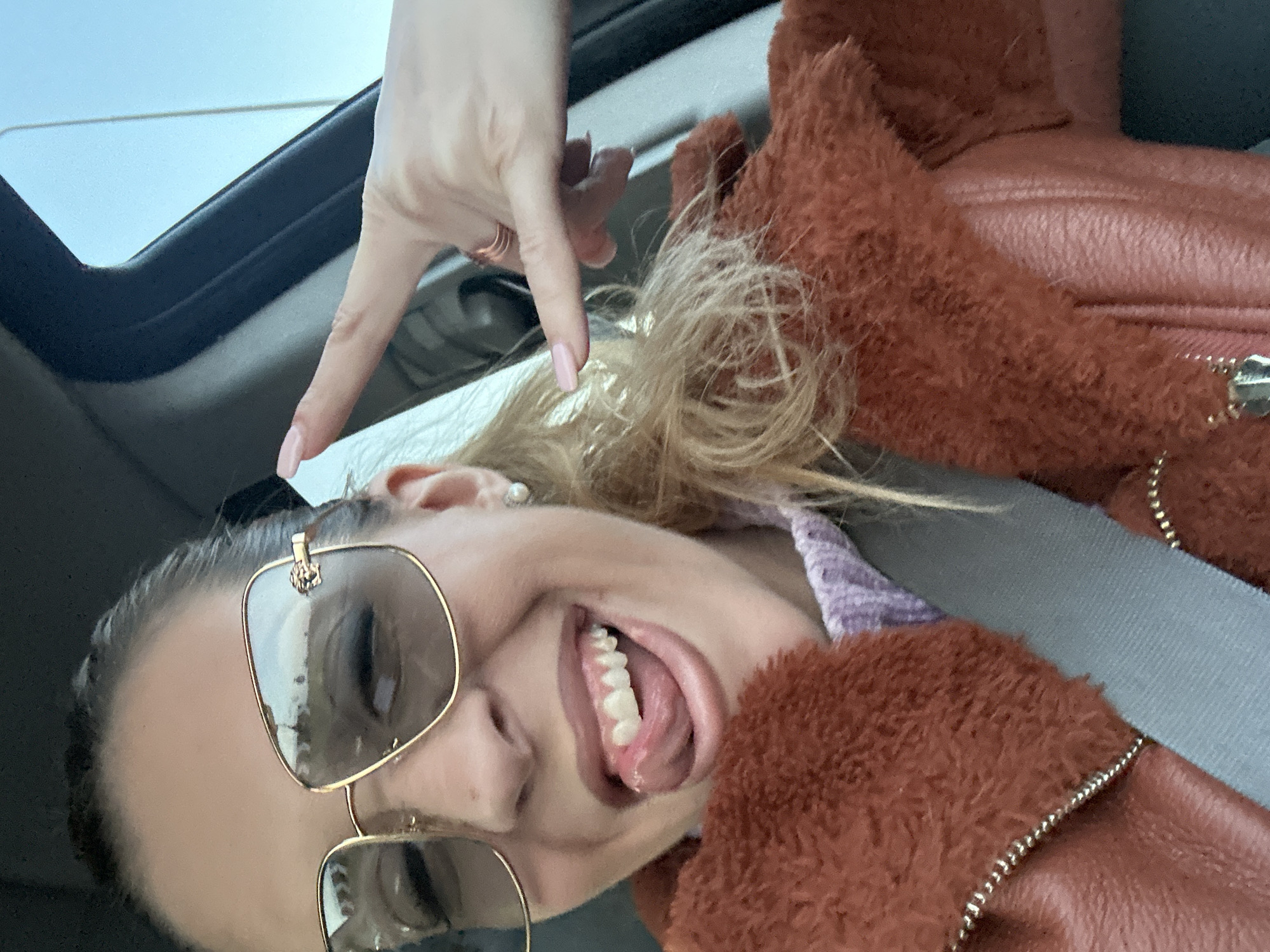 A woman wearing sunglasses and a brown jacket is in a car, possibly in Basildon, United Kingdom. She is smiling and sticking her tongue out while making a peace sign with her hand near the sunroof. She is wearing a seatbelt.