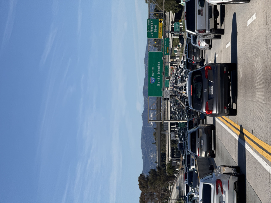 A congested multi-lane highway stretches into the distance under a clear blue sky, dotted with wispy clouds. Cars are densely packed, indicating heavy traffic. A green highway sign prominently displays "405 North Santa Monica," suggesting the location and direction of the freeway. Another sign points to "Exit 50B Jefferson Blvd." Mountainous terrain is visible in the background.