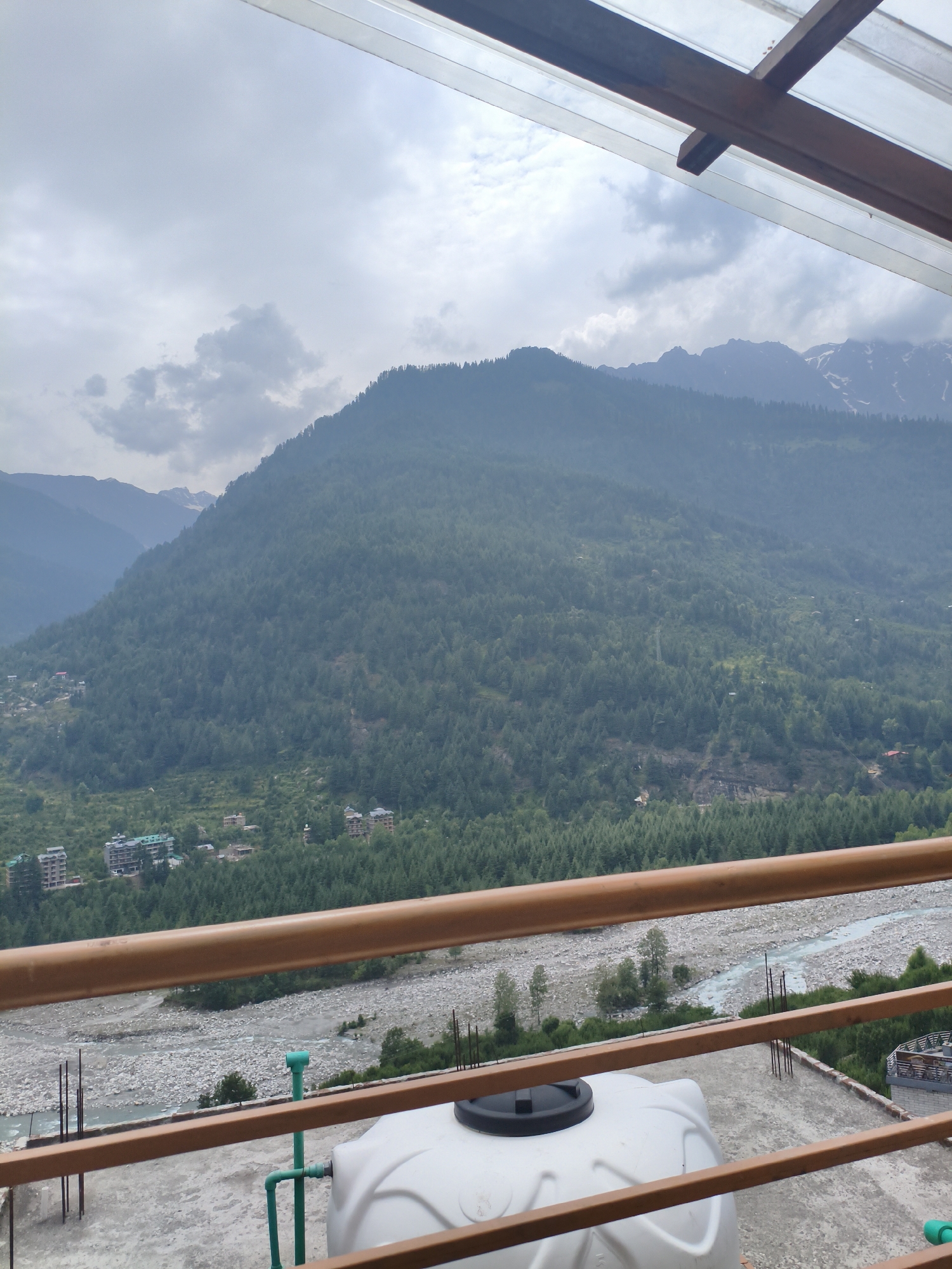 The image shows a scenic view of the valley in Vashisht, India. The foreground features a rooftop with a large white water tank and green pipes, partially obscured by brown railings. Beyond the rooftop, a rocky riverbed with a light blue river flows through the valley. The middle ground is covered in dense green coniferous forests that blanket the slopes of a large mountain. Several buildings, including apartment complexes with green roofs and smaller houses, are visible nestled within the trees and along the lower slopes of the mountain. In the background, a majestic mountain range with snow-capped peaks under a cloudy sky dominates the horizon. The overall scene depicts a natural landscape with signs of human habitation.