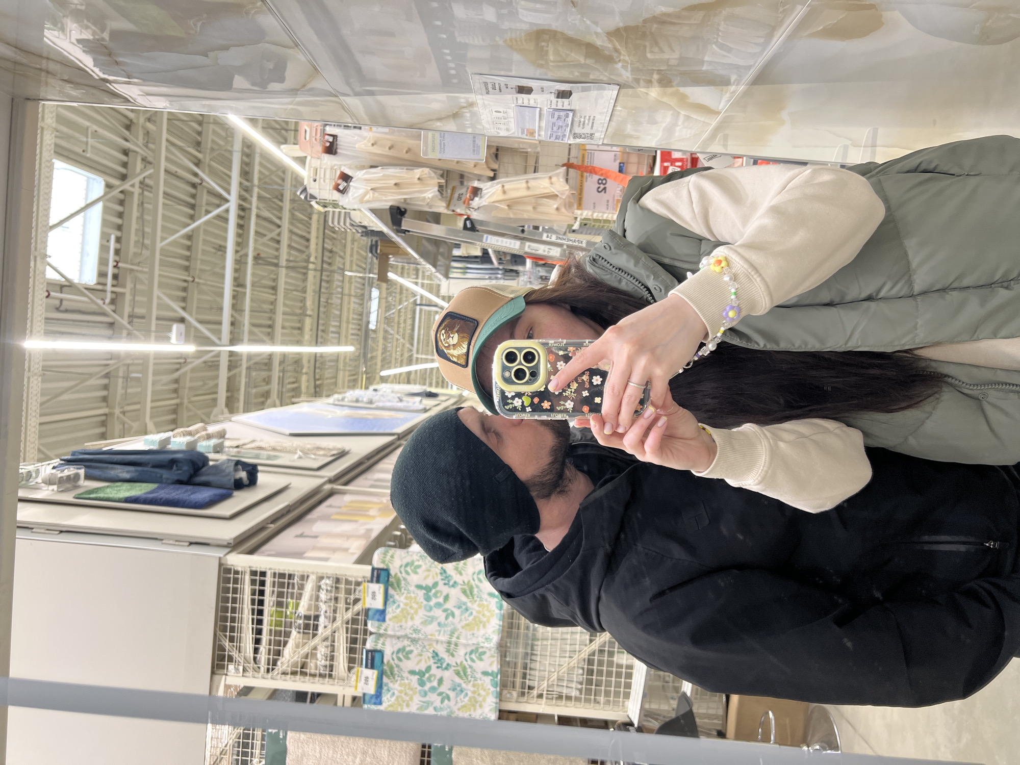 A couple is taking a selfie in a mirror, likely inside a retail store. The female is wearing a tan cap with a lion patch, a green vest, and a beaded bracelet. The male is wearing a black beanie and a black jacket. They are standing in front of shelves stocked with merchandise, including rugs and packaged items, possibly related to home decor. The ceiling is high with visible beams and lighting fixtures. I cannot tell for sure if this is the city of Kazan, Russia without additional context.