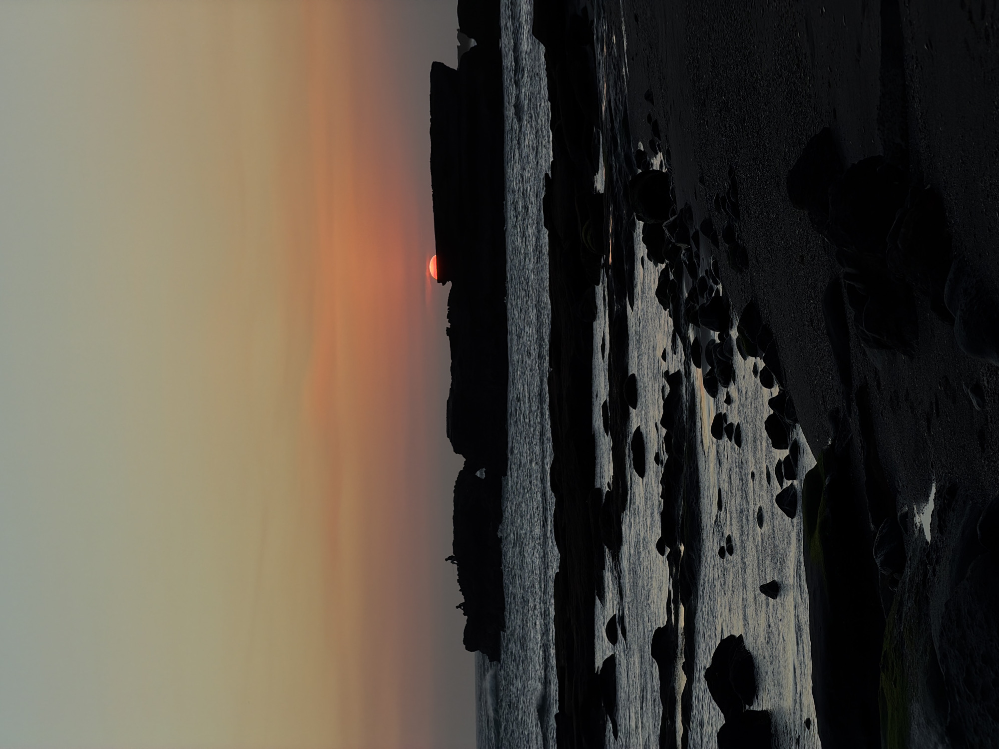 This image depicts a dramatic coastal landscape at either sunrise or sunset, originally rotated and presented here in its correct landscape orientation. The sky on the left side features a gradient of warm hues, transitioning from a soft, pale orange-yellow near the horizon to a muted grey-blue further upward. A bright, distinctive red sun is visible low on the horizon, partially obscured by a dark landmass, acting as a striking focal point.
In the mid-ground, a silhouette of a rugged coastline, likely composed of cliffs and rocky outcrops, meets the ocean. The surface of the ocean appears dark and textured, with minimal reflections, contributing to the atmospheric mood of the scene.
The right side of the frame is dominated by an expanse of dark, heavily textured rock formations and cliffs. These geological features are uneven, characterized by numerous scattered rocks and boulders. Some areas of the rock show lighter grey tones, possibly due to water erosion or mineral deposits, and faint traces of green, likely moss or algae, cling to crevices.
The lighting emphasizes deep shadows and strong silhouettes, highlighting the raw, natural beauty and ruggedness of the coastline. The low position of the sun indicates either dawn or dusk, casting a warm, enveloping glow over the sky. No people, man-made objects, or visible text are present in the image. The scene conveys a sense of stark beauty and natural grandeur, consistent with the coastal regions of Mengwi, Indonesia.