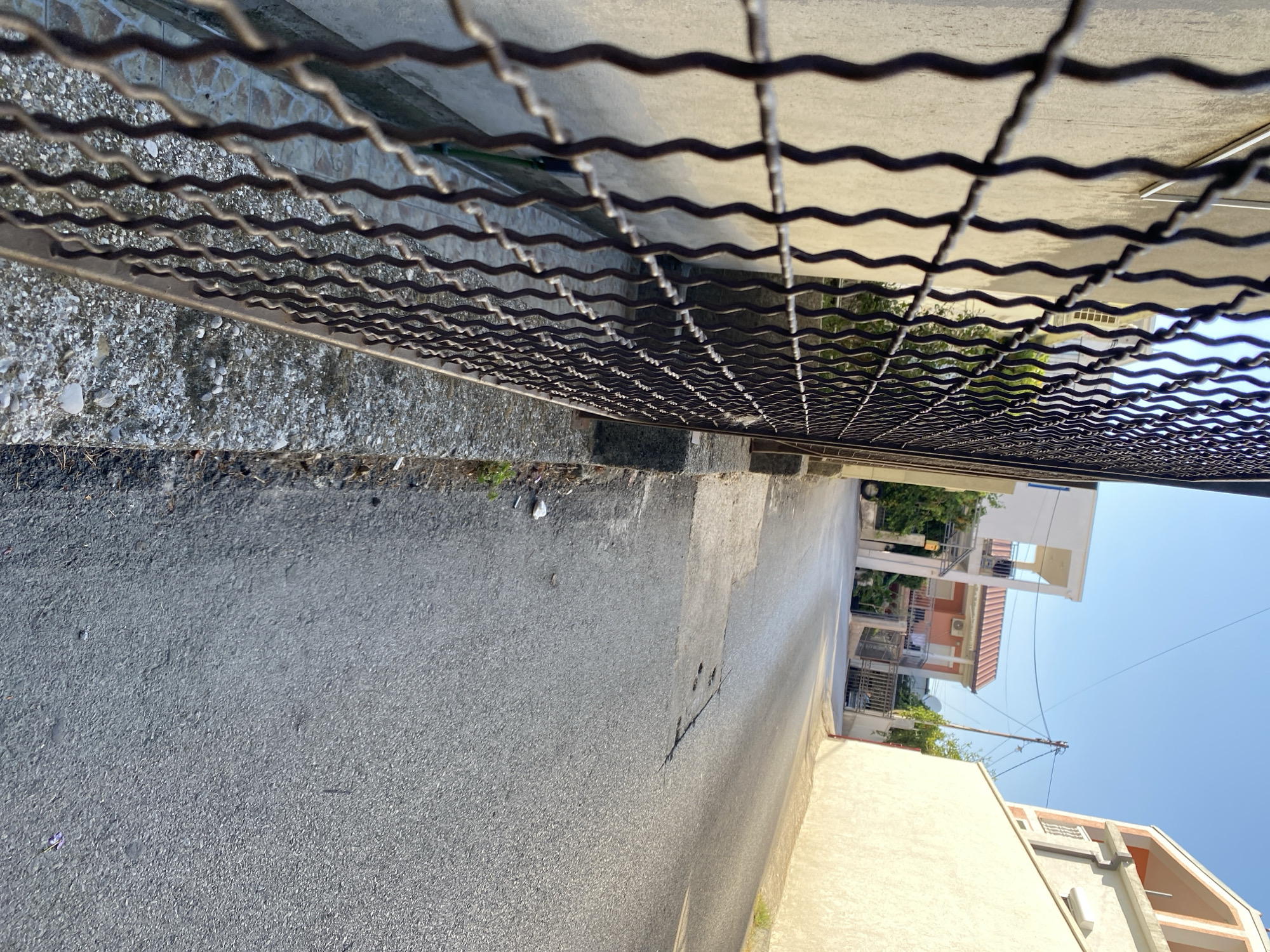 The image shows a street in Zgrade, Montenegro. A narrow, paved street runs away from the viewer and curves to the left. The street is bordered by a wall on the left and a building on the right. The wall on the left is made of concrete and has a metal fence in front of it. The building on the right is a light-colored, multi-story structure. There are several other buildings visible in the background, along with trees and a clear blue sky. The street appears to be residential, with a few cars parked along the side in the distance. The lighting suggests it is late afternoon or early morning, with long shadows cast across the scene.
