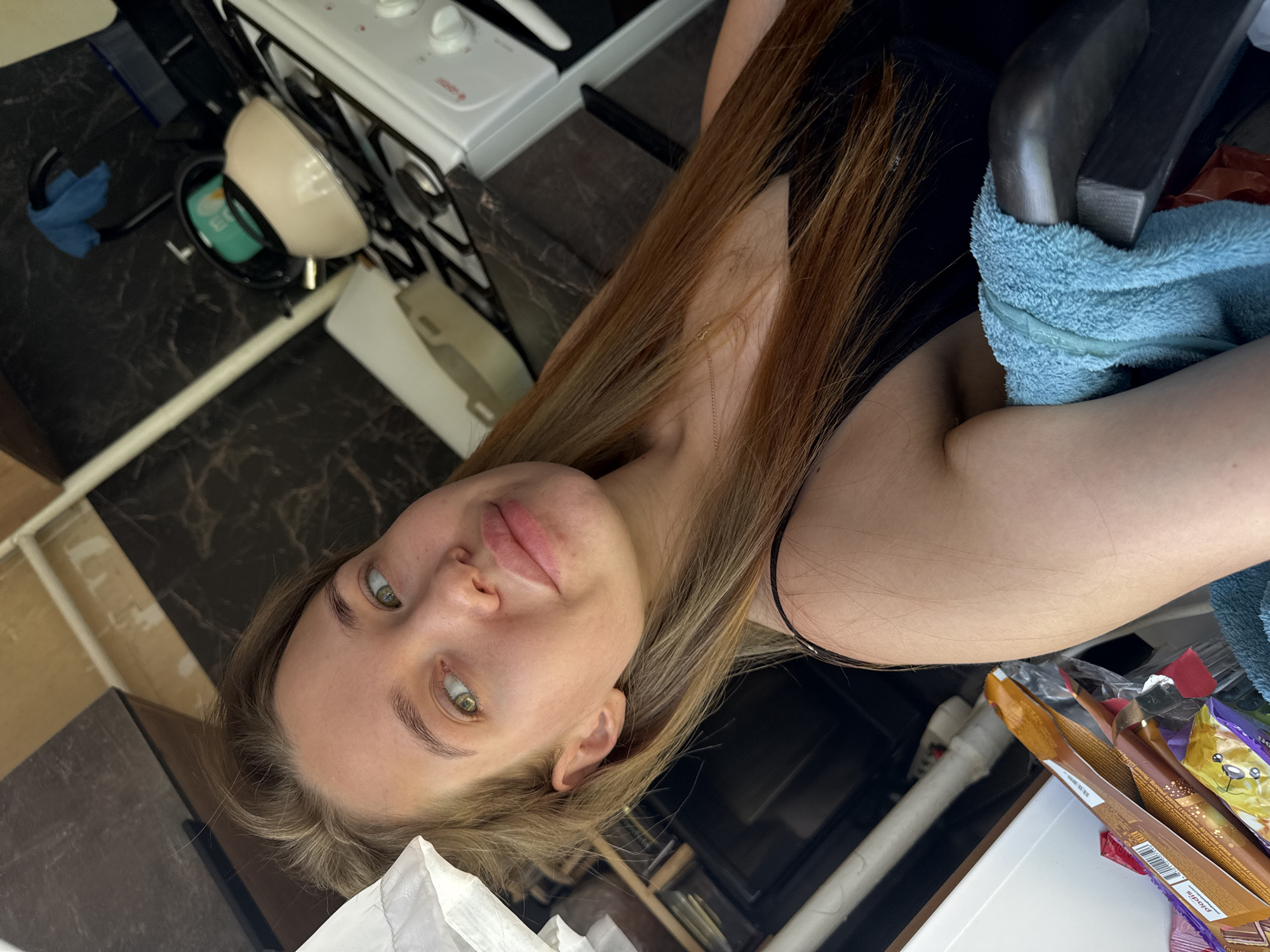 An indoor, vertically oriented image, rotated 90 degrees counter-clockwise, depicts a young woman in what appears to be a kitchen or domestic setting in Chaikovsky, Russia. The woman, the primary subject, has long, light brown hair with lighter highlights, falling over her shoulders. She looks directly at the camera with large, green eyes and a slightly parted, neutral mouth, conveying a calm expression. She is wearing a black top with thin straps, one of which is visible over her right shoulder.
The background shows various household elements. To the left of the woman's head, a white stove or cooktop with black grates and white control knobs is visible on a dark, marble-patterned countertop. A light beige bowl is placed on a black drying rack, which also holds a green container with "ECO" text and other unreadable lettering. A light blue cloth is draped over a dark object nearby.
To the right of the woman, a dark, possibly wooden, object is partly visible, with a fluffy light blue towel wrapped around it. Below this, various packaged goods and wrappers are cluttered, including a brown package displaying "zlodiz" (or similar spelling) and a barcode, as well as purple, yellow, and red packaging. A white pipe or conduit runs along the bottom left. The overall perspective is from a low angle, looking upwards at the woman. No specific activity beyond posing for the photograph is depicted.