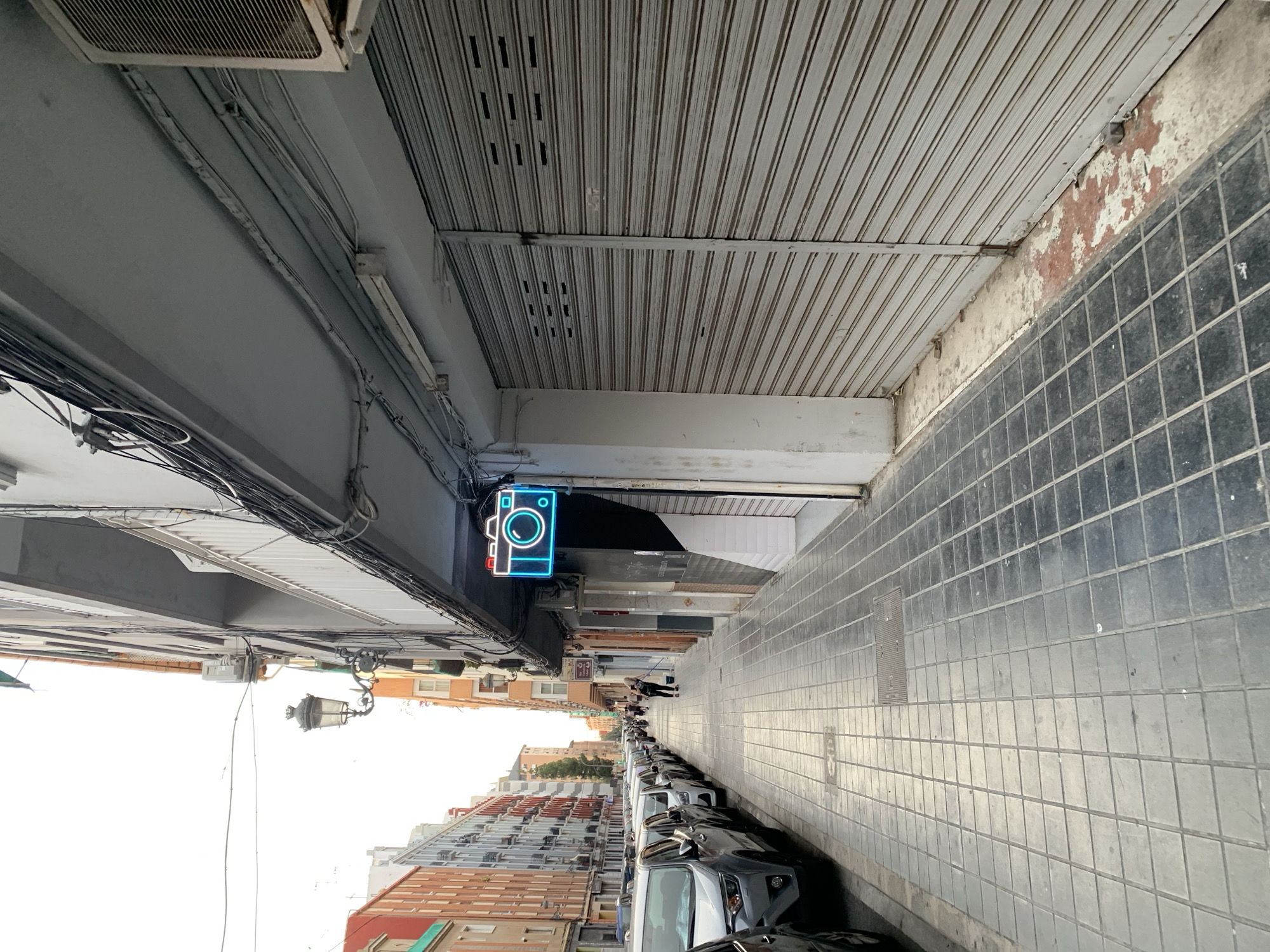 The image captures a street scene in Valencia, Spain, characterized by a row of parked cars on the left and buildings lining the right side of the street. A person is visible in the middle ground, sweeping the sidewalk. A prominent neon sign in the shape of a camera, illuminated in bright blue, is mounted above a closed storefront on the right. The architecture suggests an urban setting, with multi-story buildings and utility wires strung across the street. The pavement is tiled, and the overall atmosphere appears to be that of a quiet commercial street during the day, possibly before businesses open or after they have closed.