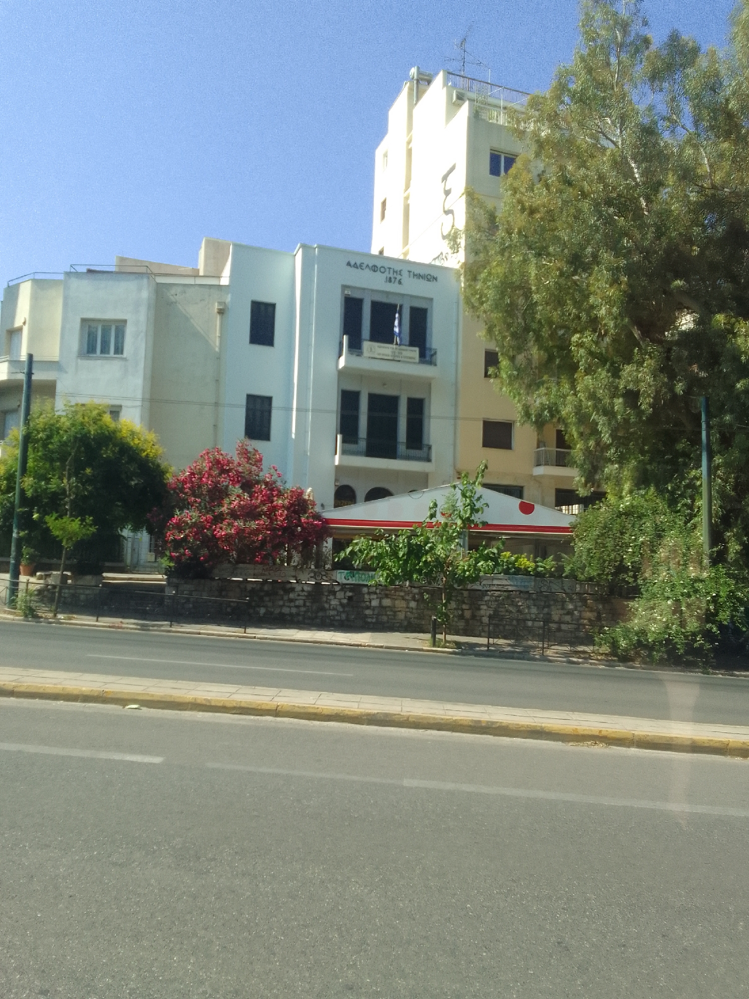 The image depicts a street scene in Athens, Greece, on a clear, sunny day. In the foreground, a multi-lane asphalt road with yellow curbing is visible. Behind the road, a low stone wall separates the sidewalk from a verdant area.
Behind the wall, several buildings are visible. The most prominent is a modern white building with a sign in Greek script that reads "ΑΔΕΛΦΟΤΗΣ ΤΗΝΙΩΝ 1876" (Hellenic Association of Tiniotes 1876). This building has multiple stories with dark-framed windows and balconies. To its left is another building, lighter in color, with a similar architectural style and a window with multiple panes. Further to the left, a glimpse of a taller, yellowish building can be seen, topped with a rooftop structure.
In front of the white building with the sign, a white awning with a red circular logo extends outwards, suggesting an outdoor seating area for a cafe or restaurant. Lush green trees and vibrant red flowering bushes add natural beauty to the scene, softening the urban architecture. A tall, slender tree with sparse foliage stands to the right, partially obscuring a tall, cream-colored building in the background. The clear blue sky indicates fair weather. The overall impression is one of a well-lit, bustling urban environment with pockets of nature. No people are clearly visible in the image.