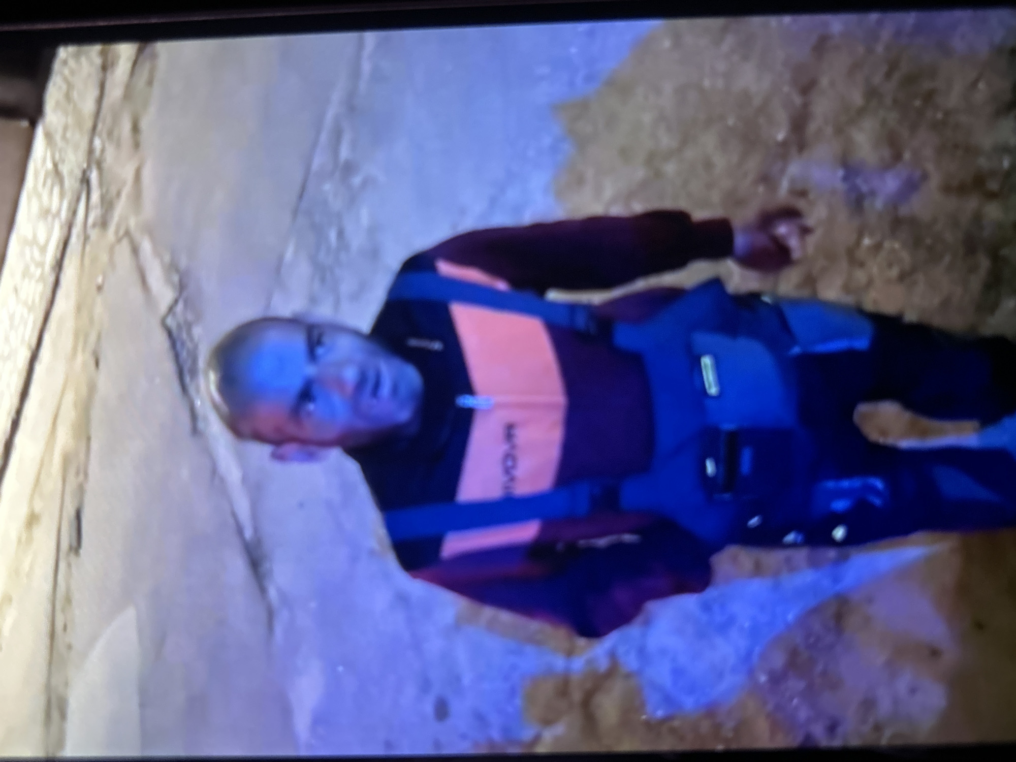 A man is depicted lying on a rough, uneven surface, illuminated by artificial blue-tinted light, suggesting an underground or enclosed natural environment. This setting is consistent with the region of Bania, Bulgaria. The man has a bald or closely shaved head and is looking towards the camera with a neutral expression. He is wearing a dark long-sleeved shirt or jacket featuring prominent orange horizontal stripes across the chest and arms. The word "VROM" is visible on one of the orange stripes. He also has a dark harness-like apparatus secured around his waist and legs, commonly used for activities such as caving, climbing, or safety. The ground appears to be a mix of light-colored, textured rock or earth and darker, possibly damp or muddy, areas. His arms are extended to his sides. The image portrays an individual engaged in an activity within a subterranean landscape.