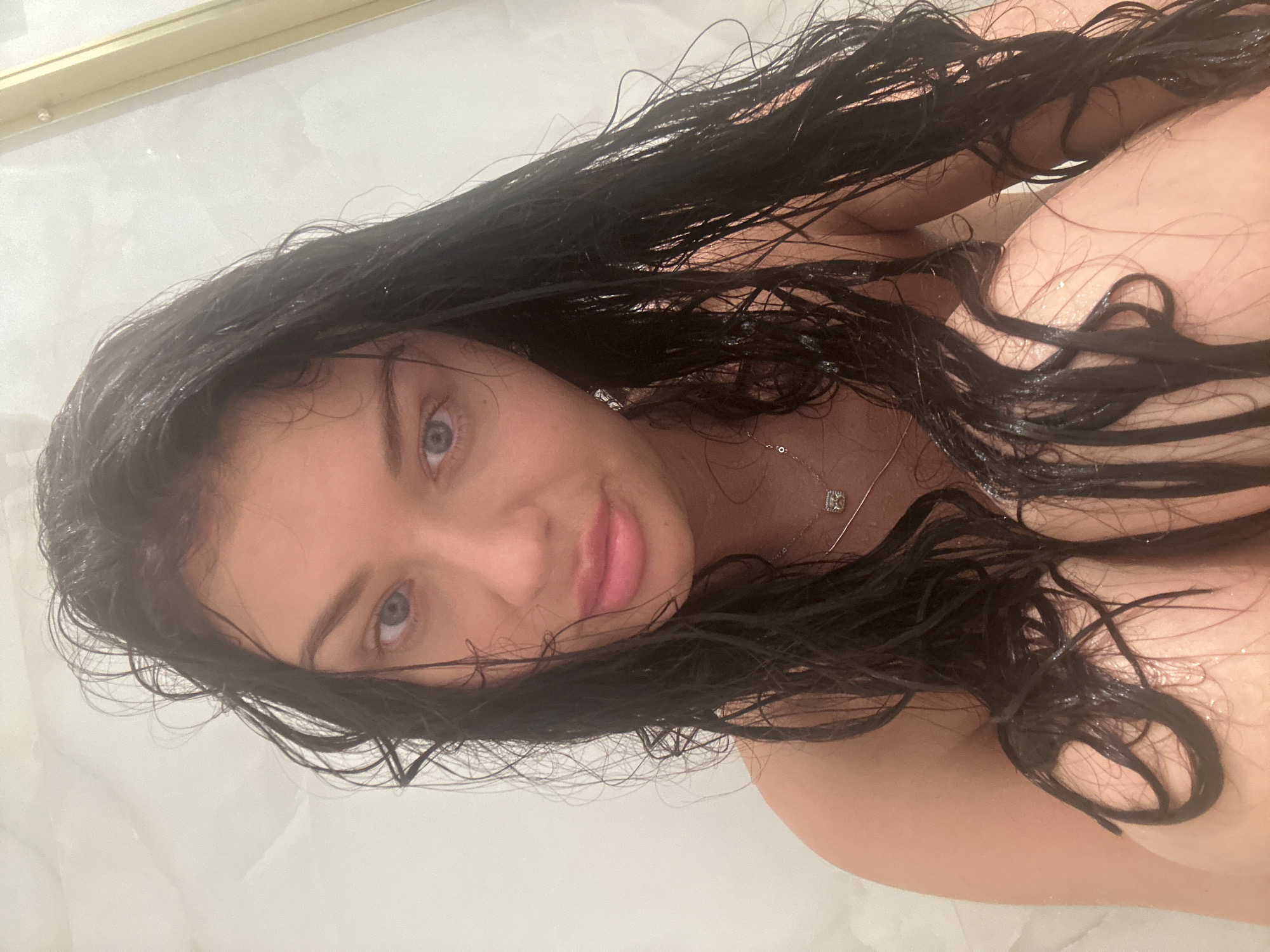 A close-up, rotated portrait features a woman with long, dark, wet hair splayed around her. She has striking blue eyes and full, pink lips, looking directly at the viewer with a calm and engaging expression. Her hair is visibly damp, with water droplets clinging to the strands and her skin. A delicate silver-toned necklace with a small, square pendant adorns her neck. The background is a light, uniform color, suggesting an indoor, possibly tiled or glass shower or bath environment. Her body is partially visible, wet, and appears to be in a bathing setting. The image captures an intimate moment, likely a selfie. No specific time of day or weather is discernible. No visible text is present. The context provided is Gavrilkovo, Russia.