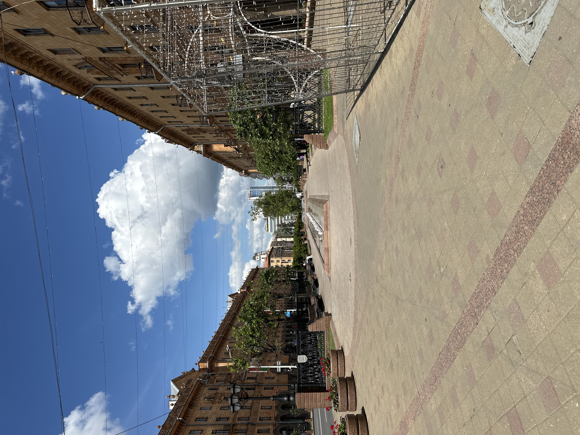 The image depicts a wide pedestrian walkway in Minsk, Belarus, featuring the characteristic Soviet-era architecture. The buildings flanking the walkway are tall, multi-story structures with light-colored stone facades, adorned with decorative cornices and small balconies. The sky is blue with scattered white clouds, suggesting a clear day.
In the foreground, the walkway is paved with square tiles in shades of beige and reddish-brown, bordered by a central strip of pinkish granite. To the left, a black lamppost stands tall, accompanied by circular planters filled with red flowers. Further along the walkway, benches are visible, providing seating for pedestrians. A black metal fence separates the walkway from the grassy area and trees.
In the middle ground, a fountain is visible, with water jets spraying upwards. The walkway extends into the distance, lined with trees and more buildings. In the far distance, modern skyscrapers and older architectural elements blend together, indicating the city's evolution.
On the right side of the image, a large, unfinished metal structure, possibly a part of an outdoor installation or temporary decoration, occupies a significant portion of the frame. The structure is made of wire mesh and appears to be lit by numerous small lights.
There are a few people visible in the image, including one individual sitting on a bench in the middle ground and another walking in the distance. The overall scene conveys a sense of urban life on a sunny day in a European city.