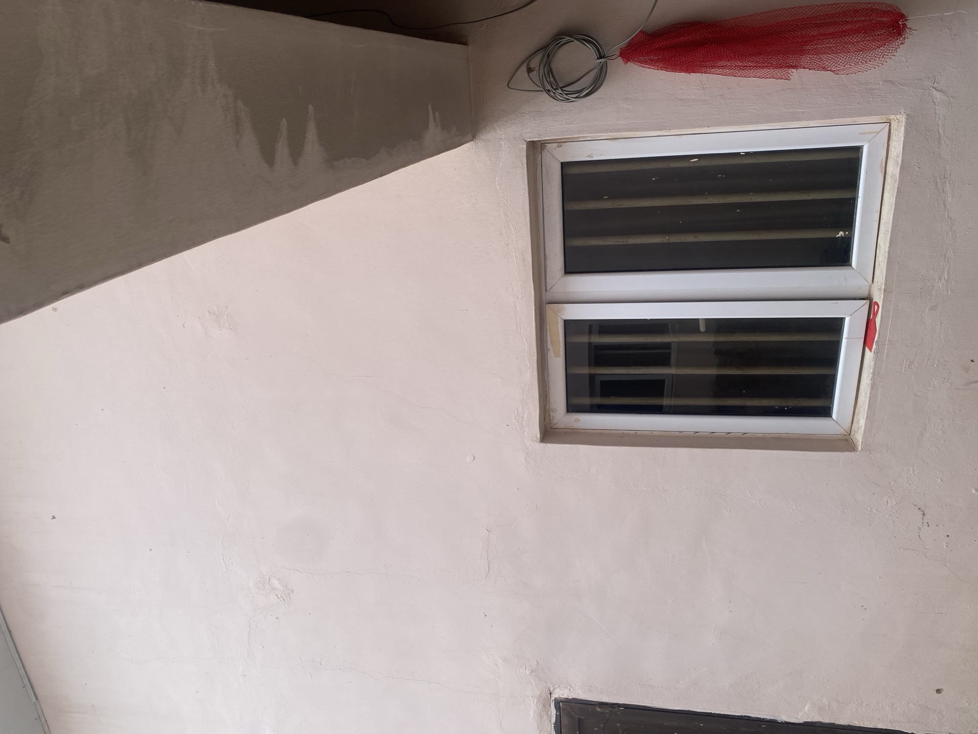 This media file captures an outdoor view of a building under construction or renovation. The scene is dominated by a pale pink, textured wall that shows signs of uneven plastering, with some small cracks and darker marks. To the right of the wall, two horizontal windows with white frames are visible. These windows have dark louvers or blinds, obscuring the view inside. Above the windows, a coiled electrical wire hangs loosely, and a bright red mesh bag is tied to a wire, protruding from the top right corner of the frame. The upper left portion of the image features a contrasting grey surface, possibly a ceiling or overhang, which also appears to have a rough, unfinished texture. The lighting suggests it is daytime, and the location cues, such as the building materials and the outdoor setting, point to a construction or renovation site, possibly in Dumnga, Nigeria. There is no discernible text in the image, and no people or specific activities are depicted. The overall impression is one of an unfinished or developing space.